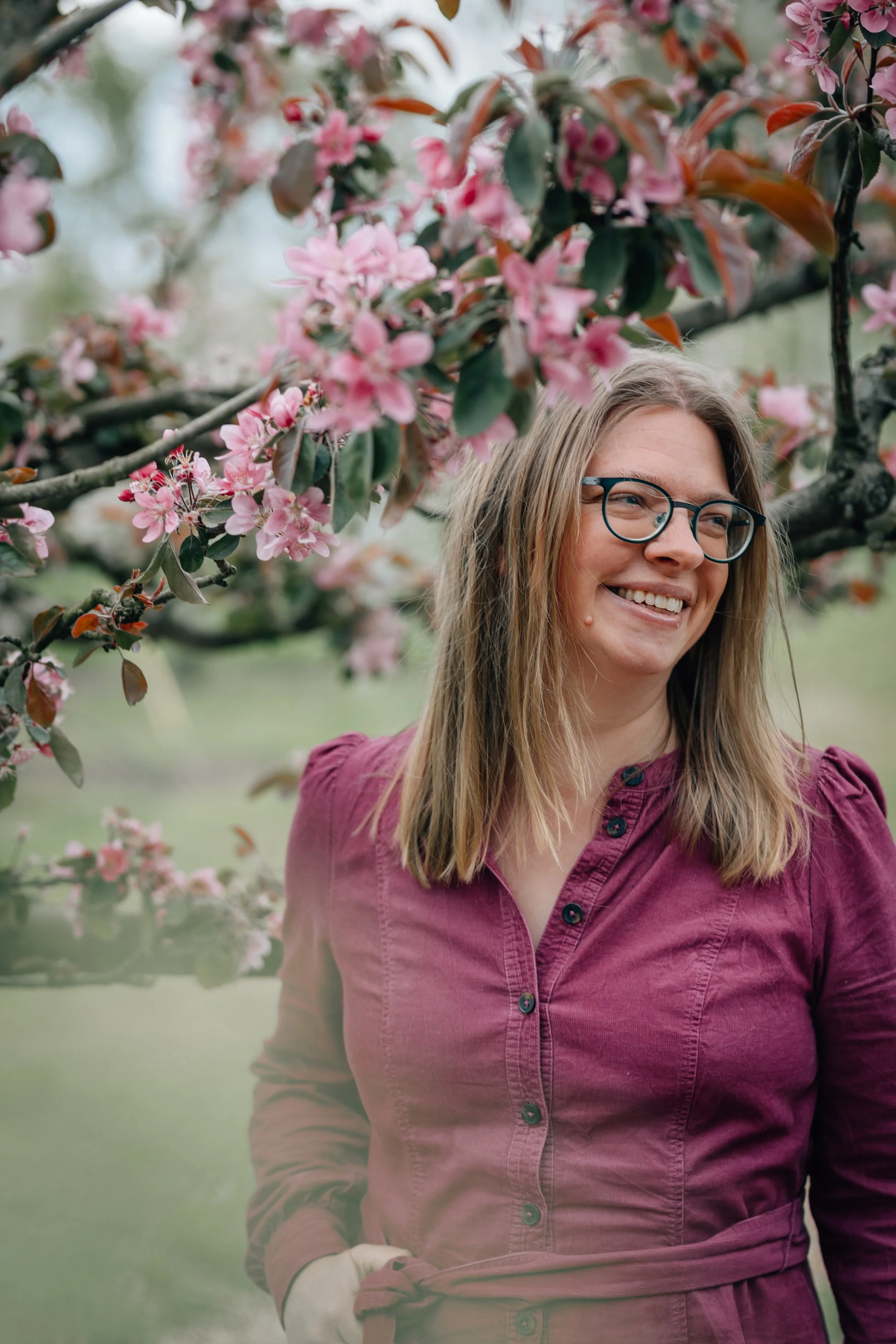 Eine Frau mit, braunem Haar und einem Lila-Kragen-Shirt steht vor blühenden rosa Kirschblütenbäumen im Freien, lächelt und schaut nach rechts.