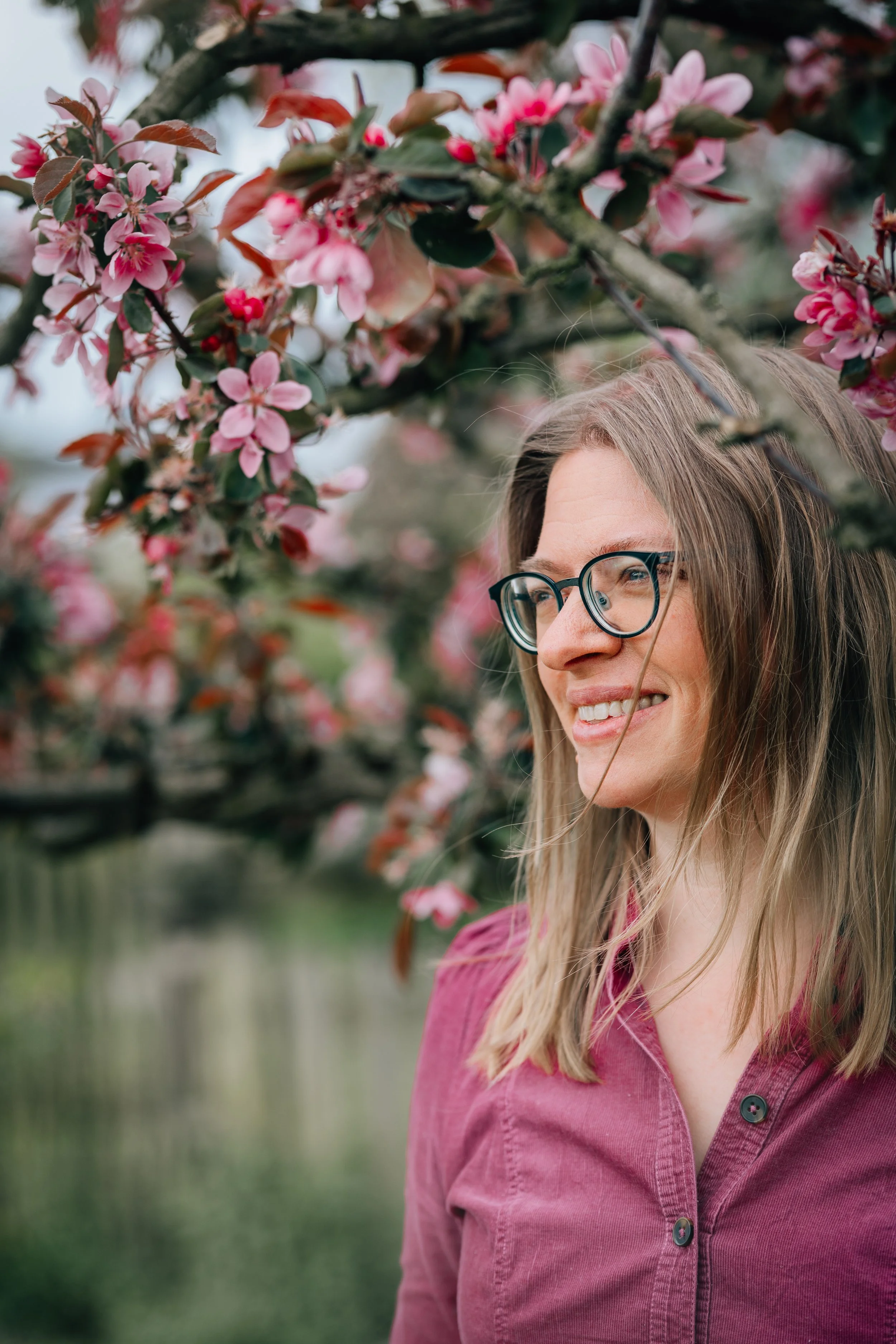 Eine lachende Frau mit Brille steht vor blühenden Kirschblüten auf einem Baum im Freien.
