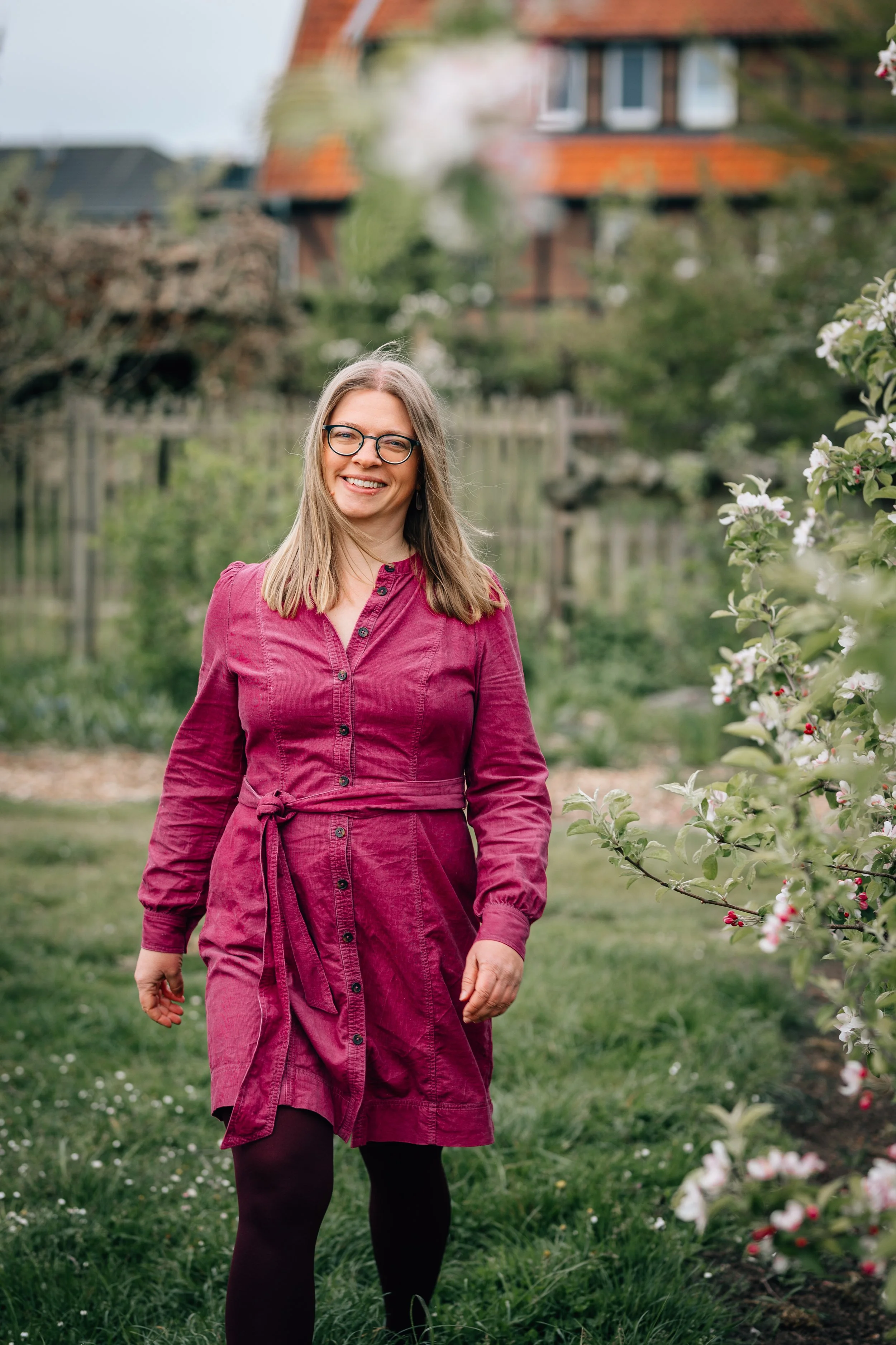 Eine lächelnde Frau mit Brille trägt ein rosa Kleid und spaziert im Garten, umgeben von blühenden Bäumen und einem Haus im Hintergrund.