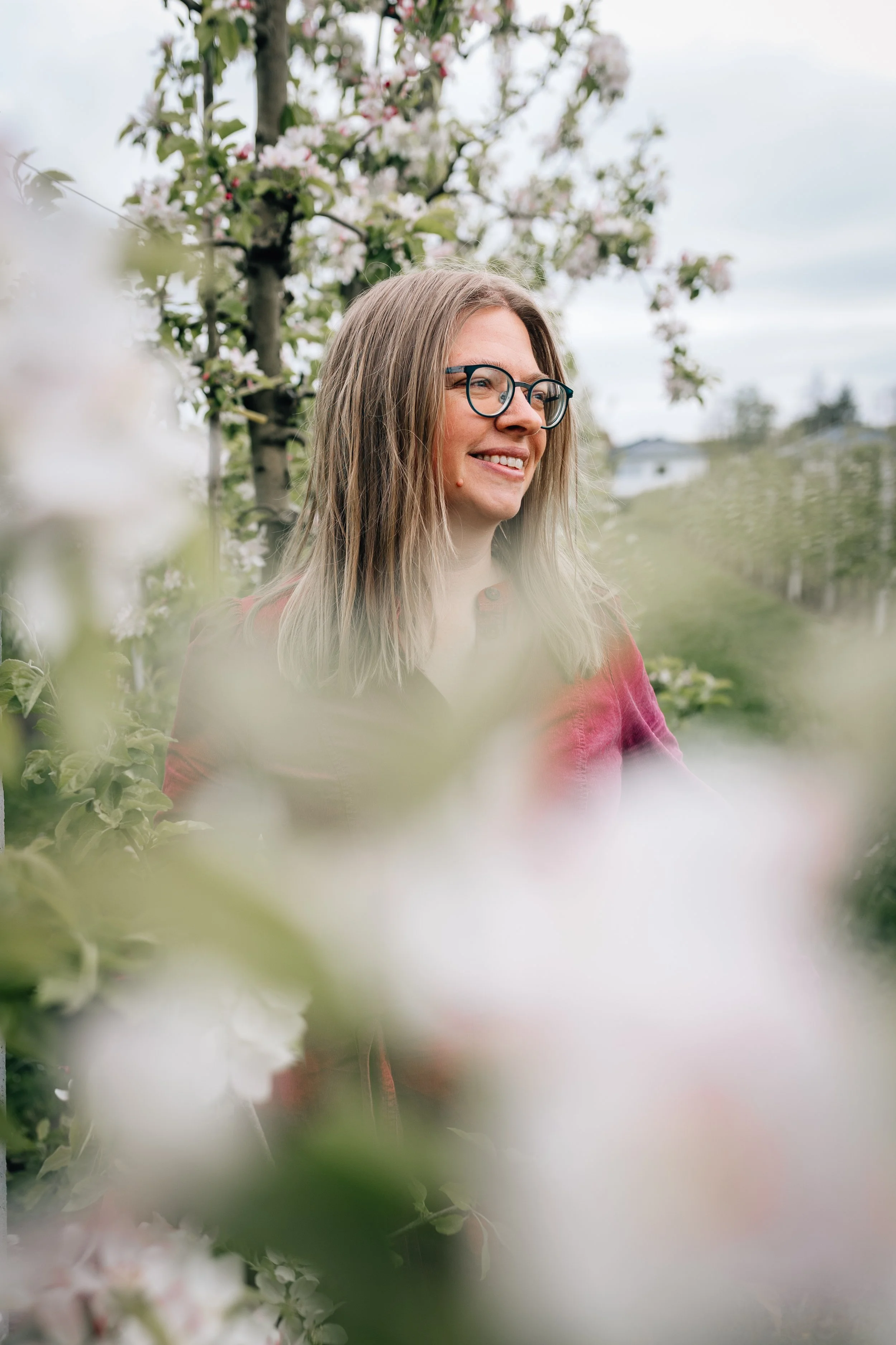 Junge Frau mit Brille und langen Haaren in einem blühenden Obstgarten, Blick nach rechts, lächelnd.