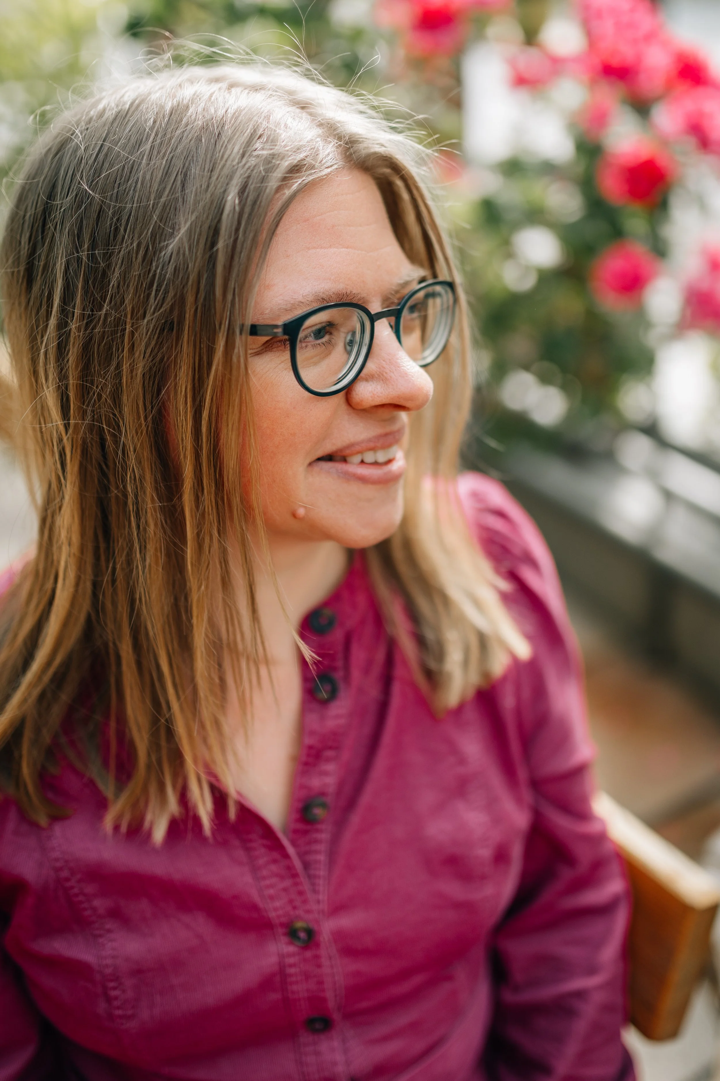 Nahaufnahme einer Frau mit langen blonden Haaren, trägt schwarze Brille, lächelt leicht, im Hintergrund sind rosa Blumen und grüne Blätter.
