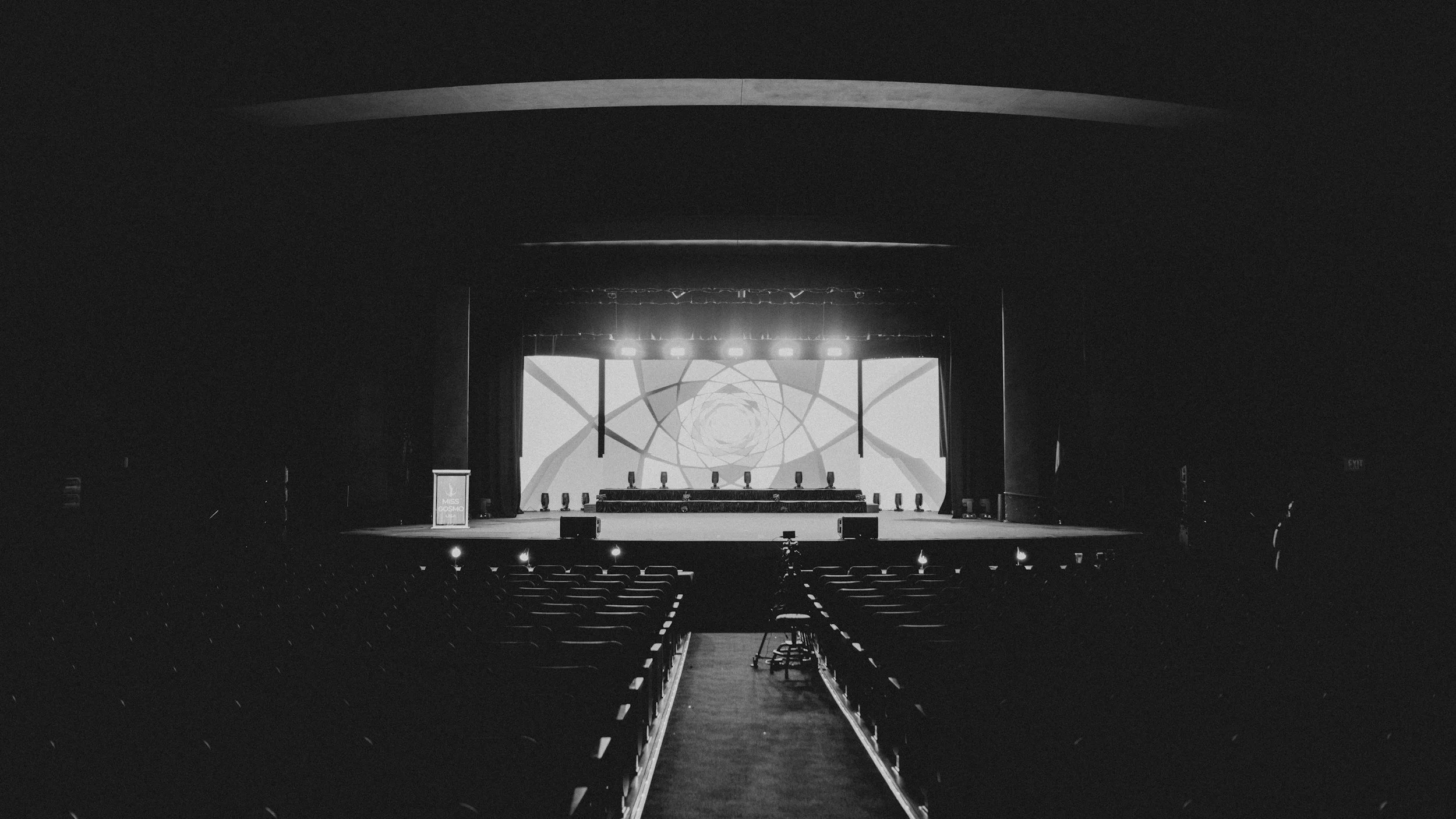 Empty stage with a large screen displaying geometric patterns, set in a dark auditorium with seats and some stage lighting.