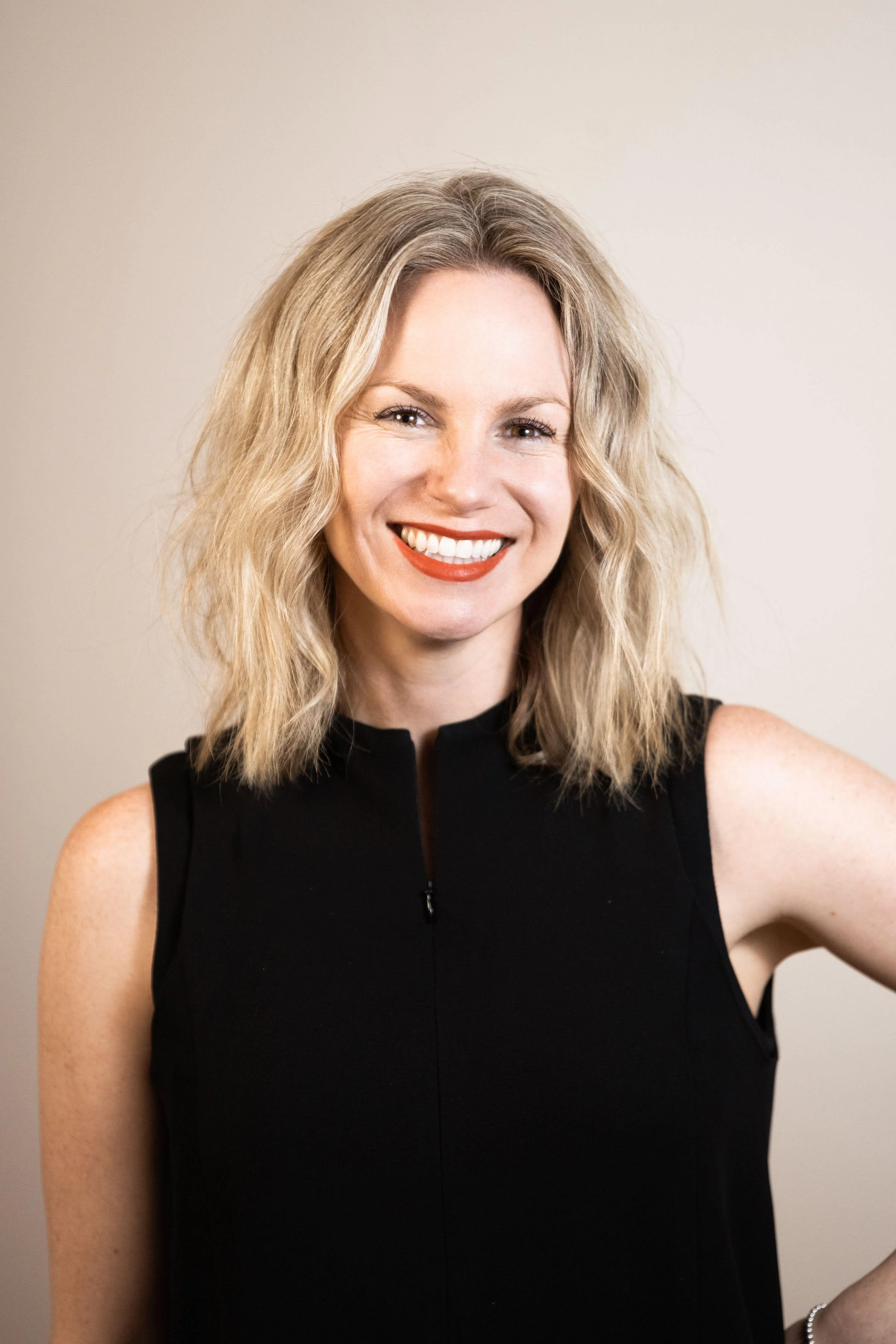 A woman with shoulder-length wavy blonde hair smiling, wearing a black sleeveless dress and a silver bracelet, standing against a plain beige wall.