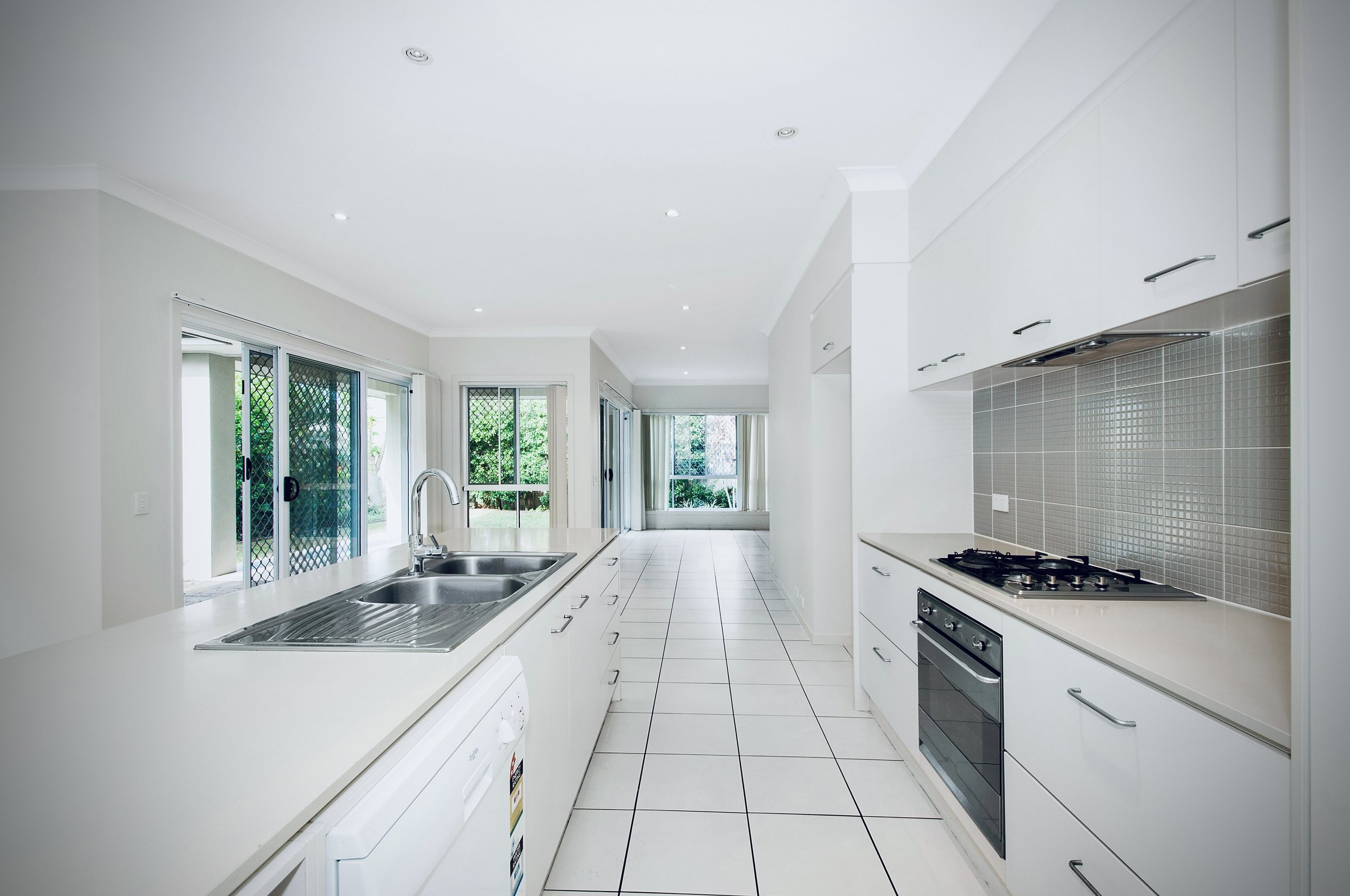 Bright, modern kitchen with white cabinets, tiled backsplash, stainless steel sink, gas stove, and large windows with sliding glass doors leading outside.