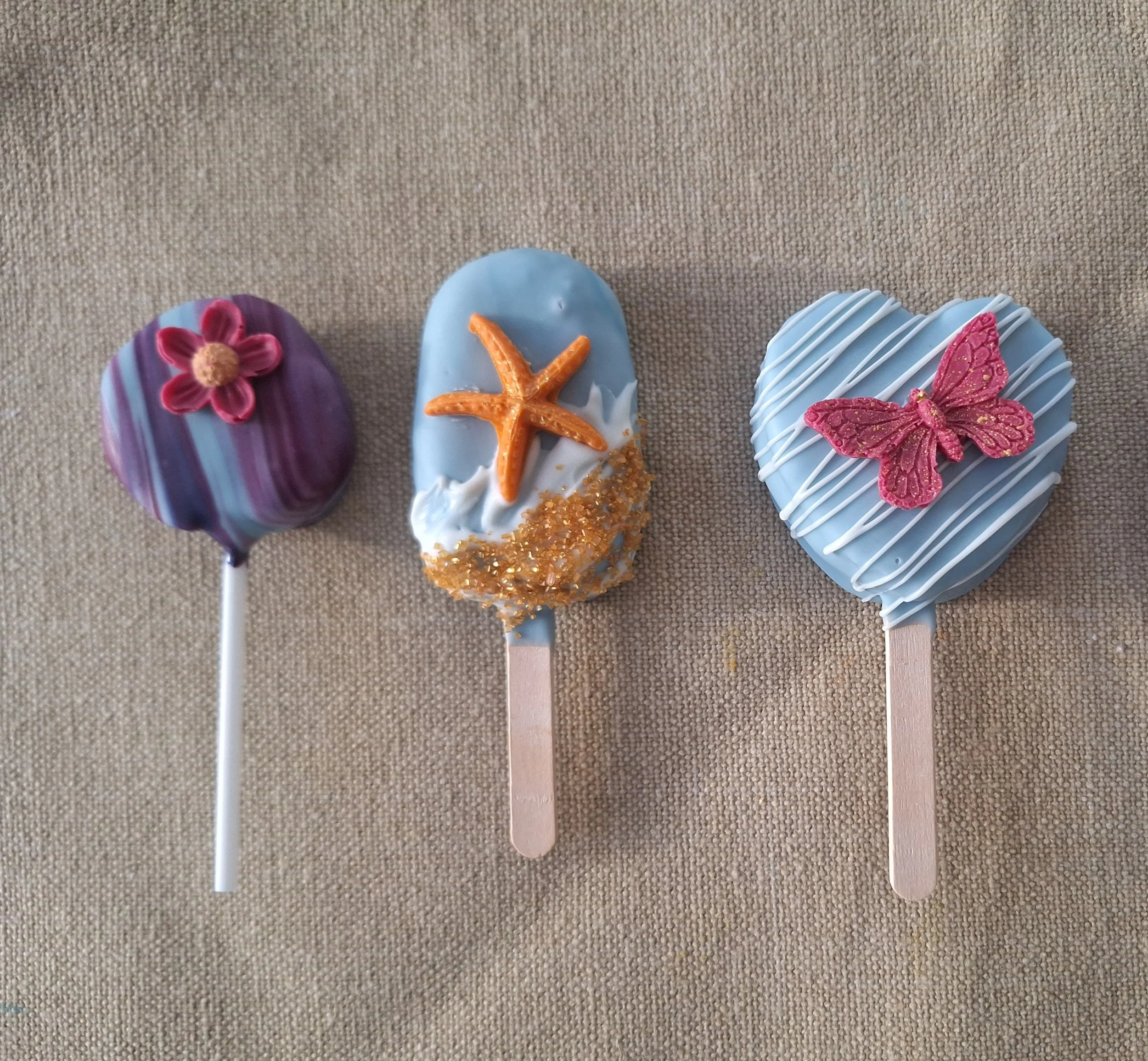 Four decorated cakesicles and cake pops with different floral and ocean-themed designs on a beige fabric background.