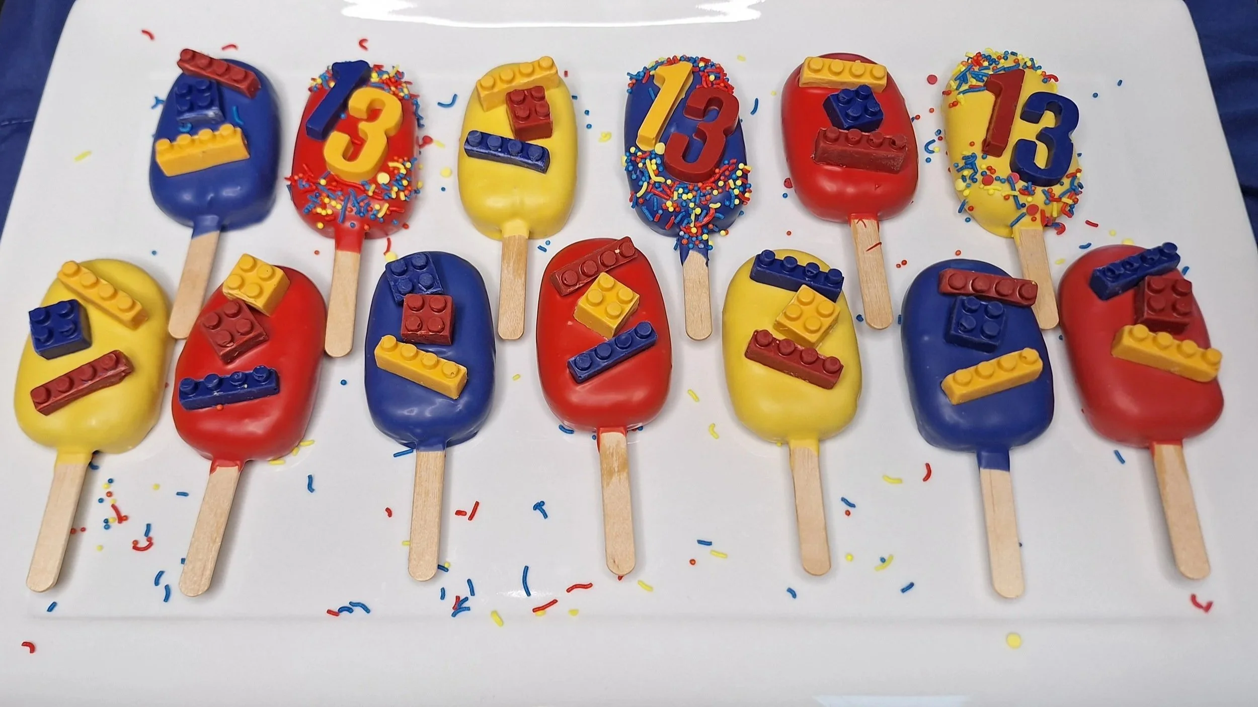 Colorful cakesicles decorated with sprinkles and Lego-themed edible decorations, arranged in three rows on a white surface.
