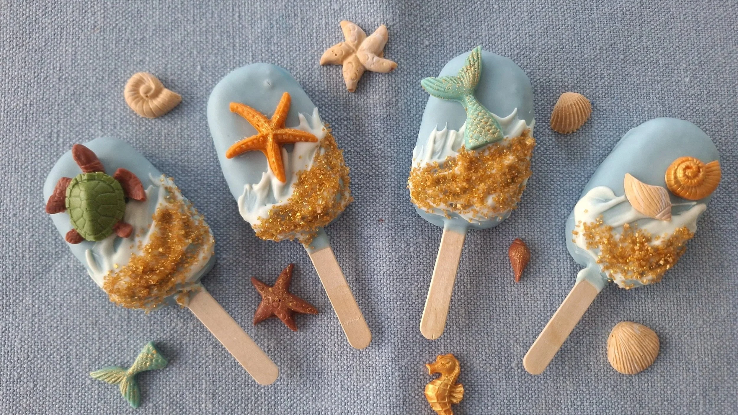Four blue cakesicles decorated with sea-themed edible toppers, including starfish, mermaid tails, a turtle, seashells, and sand-like sprinkles on a gray textured surface, with small seashells and starfish around them.