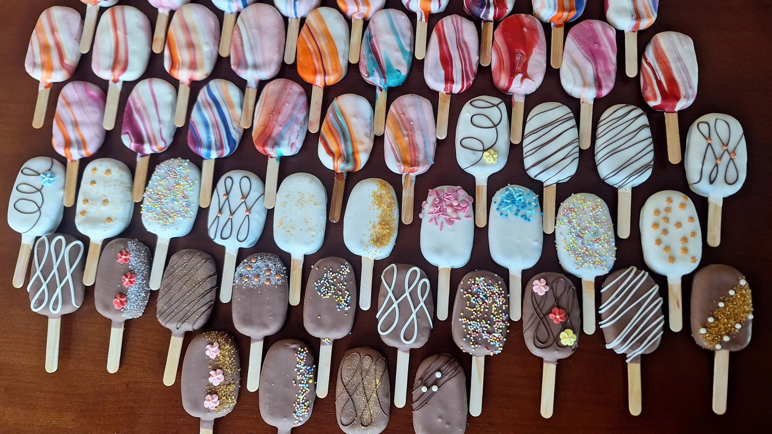 Assorted decorated cakesicles, some with colorful stripes, sprinkles, and chocolate coatings, arranged on a dark surface.