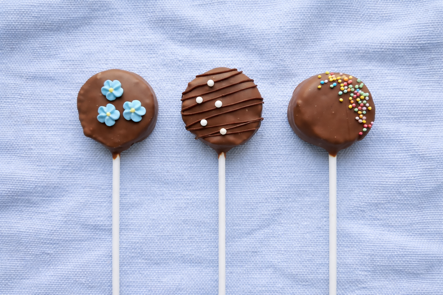 Three chocolate-covered cake pops decorated with colorful sprinkles and flowers on a light blue textured background.