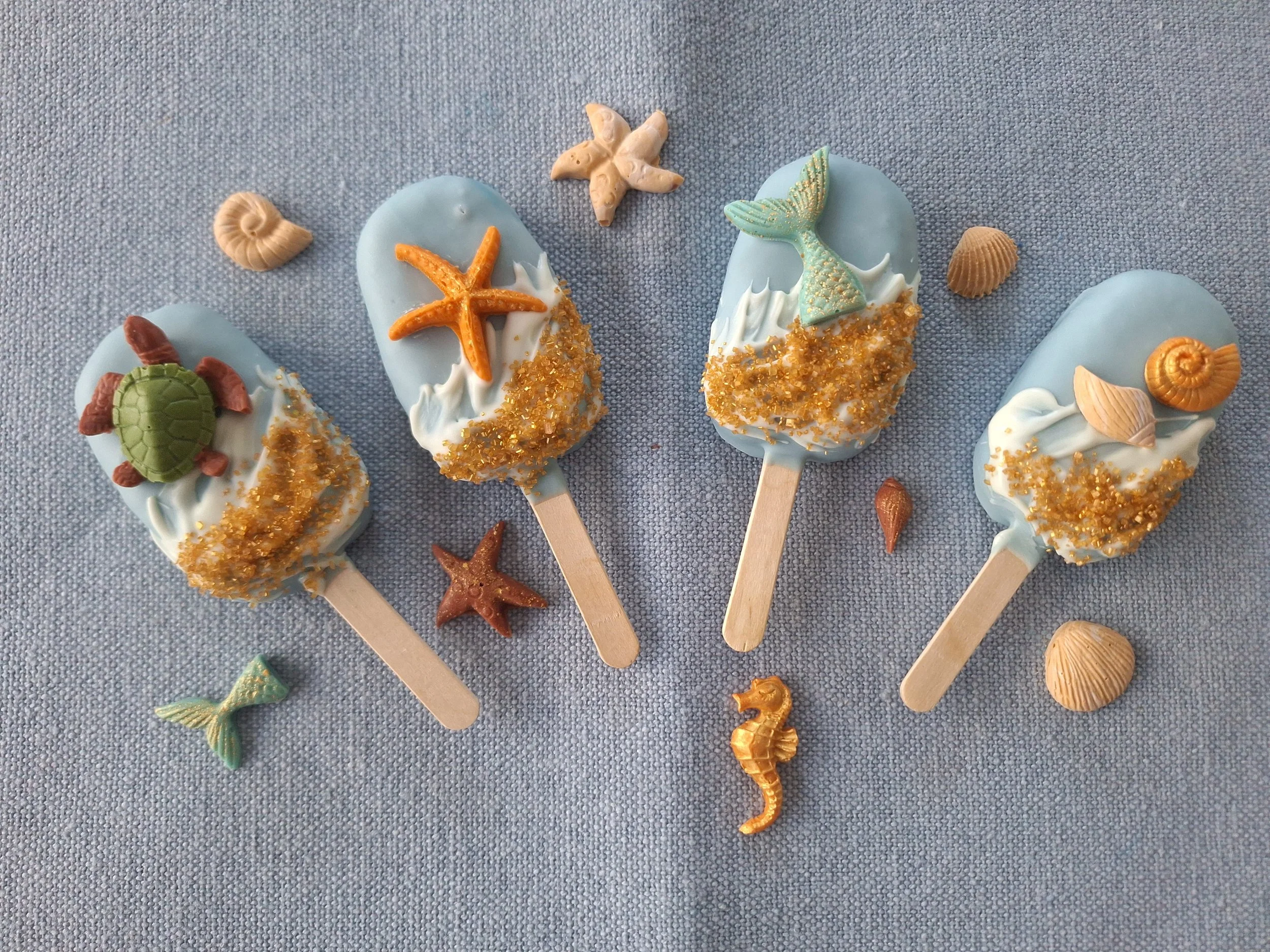 Light blue cakesicles decorated with sea-themed candies such as starfish, shells, a mermaid tail, a turtle, and a seahorse on a gray textured surface.
