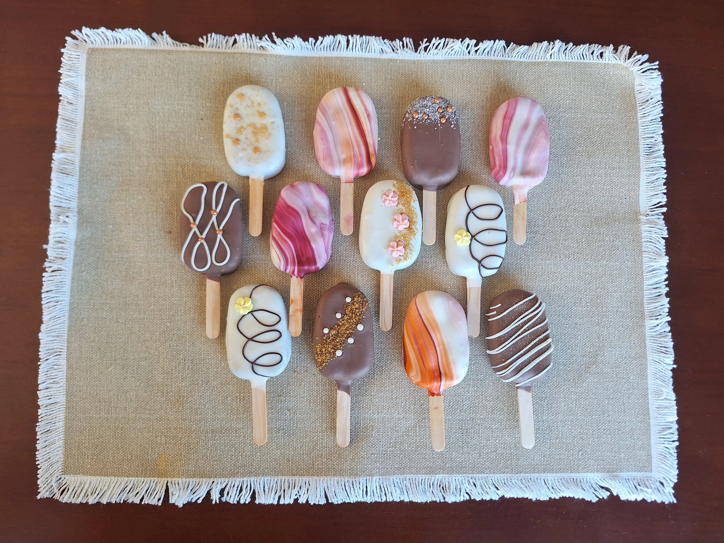 Assorted decorated cakesicles on a beige fringed cloth on a dark wooden table.