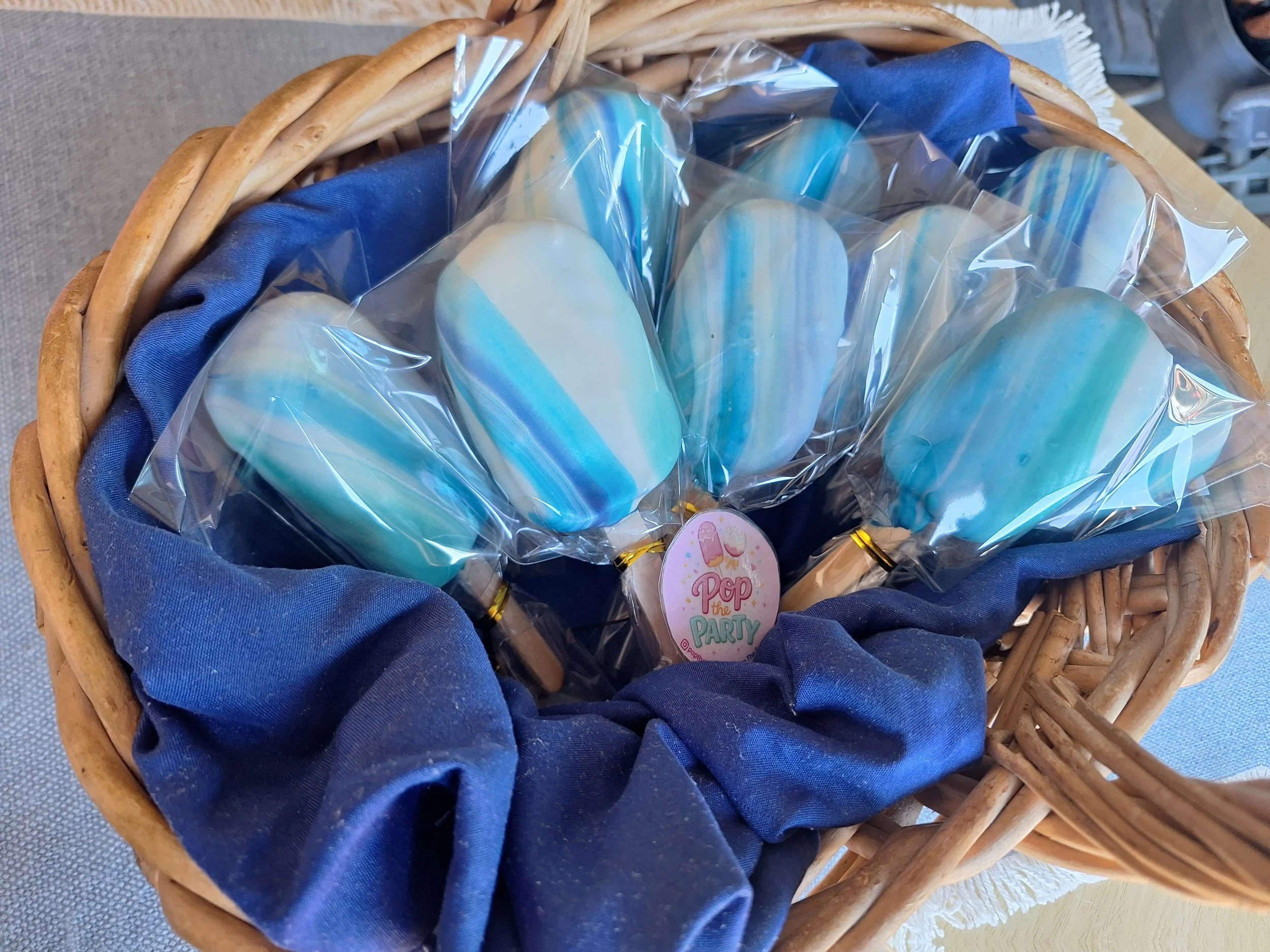 A wicker basket lined with blue fabric holds wrapped cakesicles decorated with blue and white stripes, and a sticker reading 'Pop the Party'.