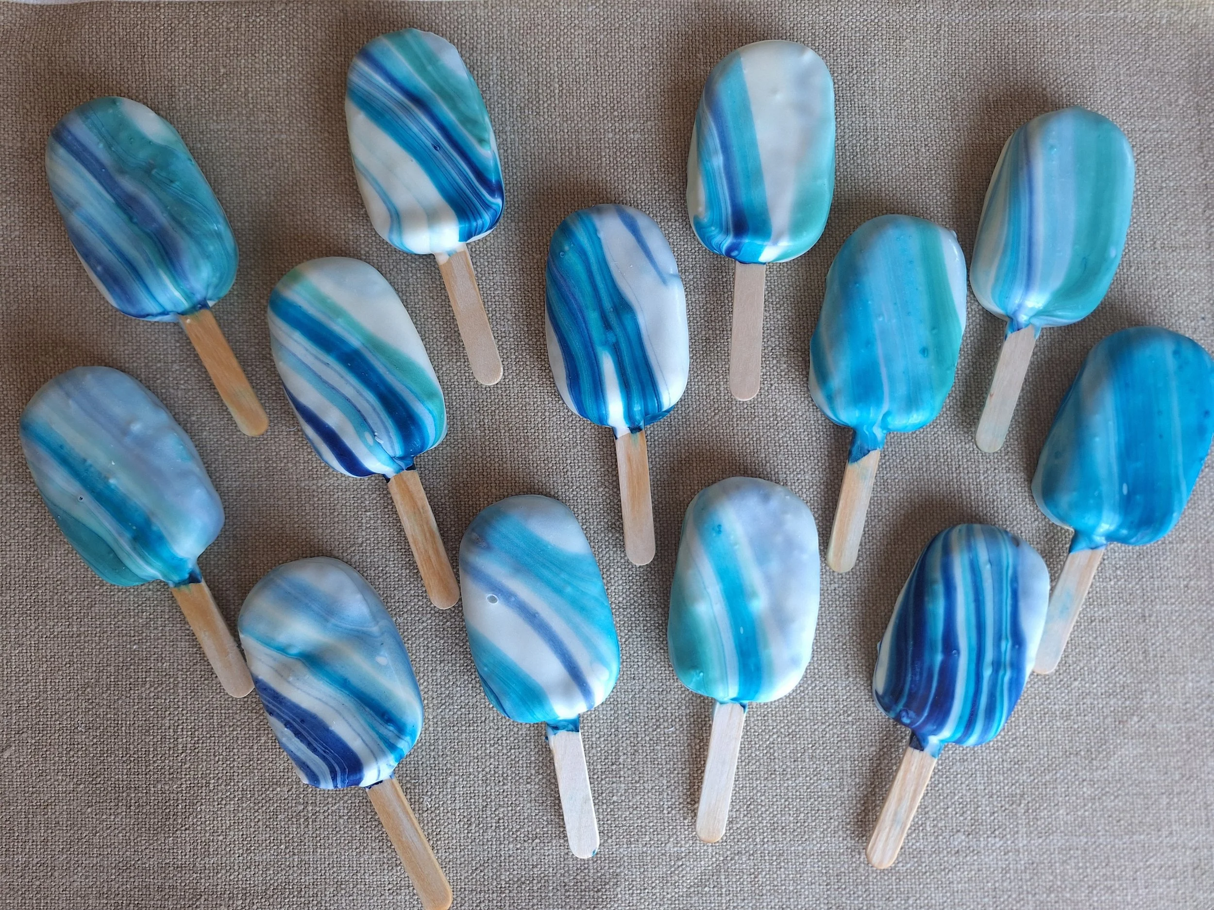 Multiple blue, white, and teal marbled cakesicles with wooden sticks arranged on a beige fabric background.