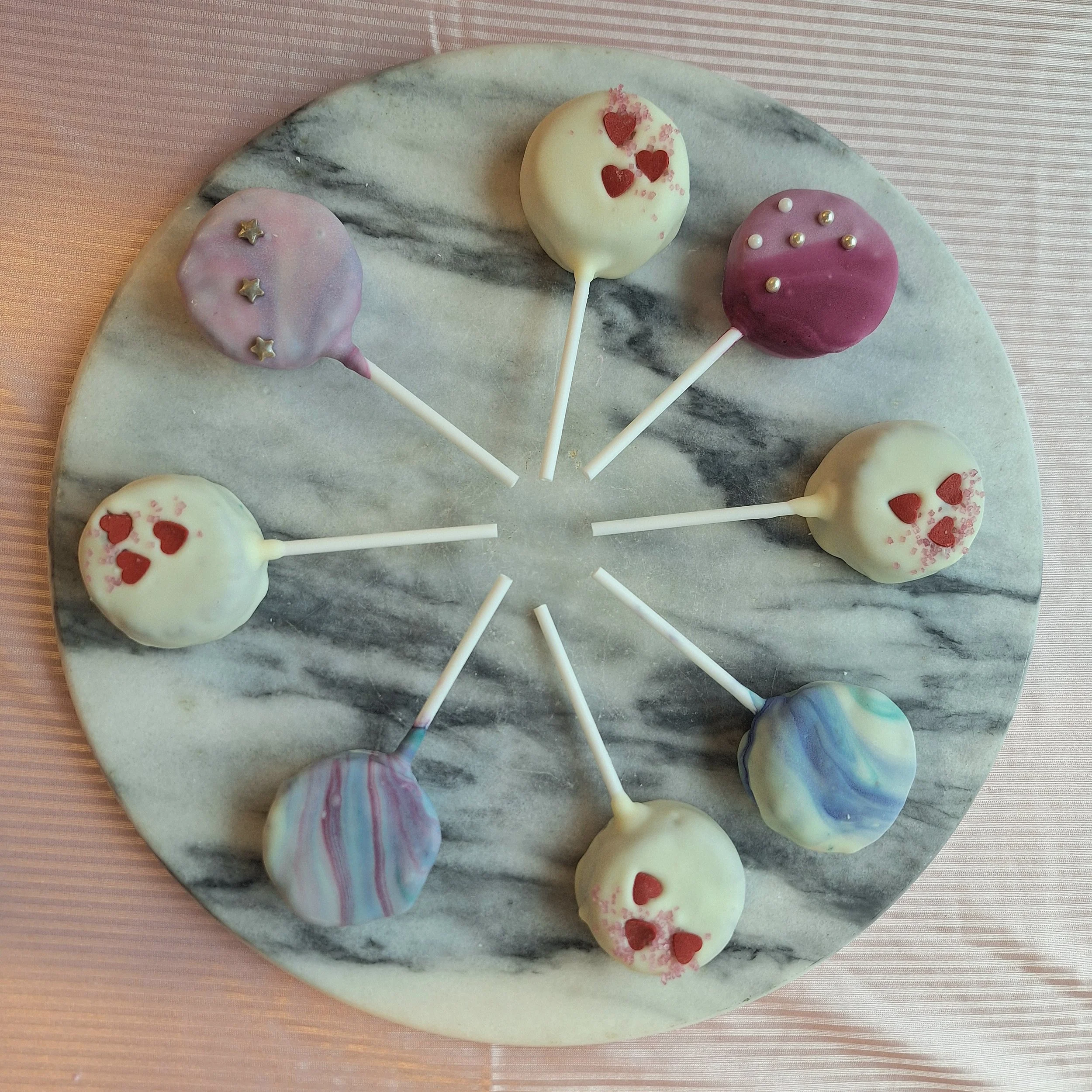 Marble surface with decorated cake pops in white, pink, and rainbow colors, some with red heart and star toppings, on a pink striped tablecloth.
