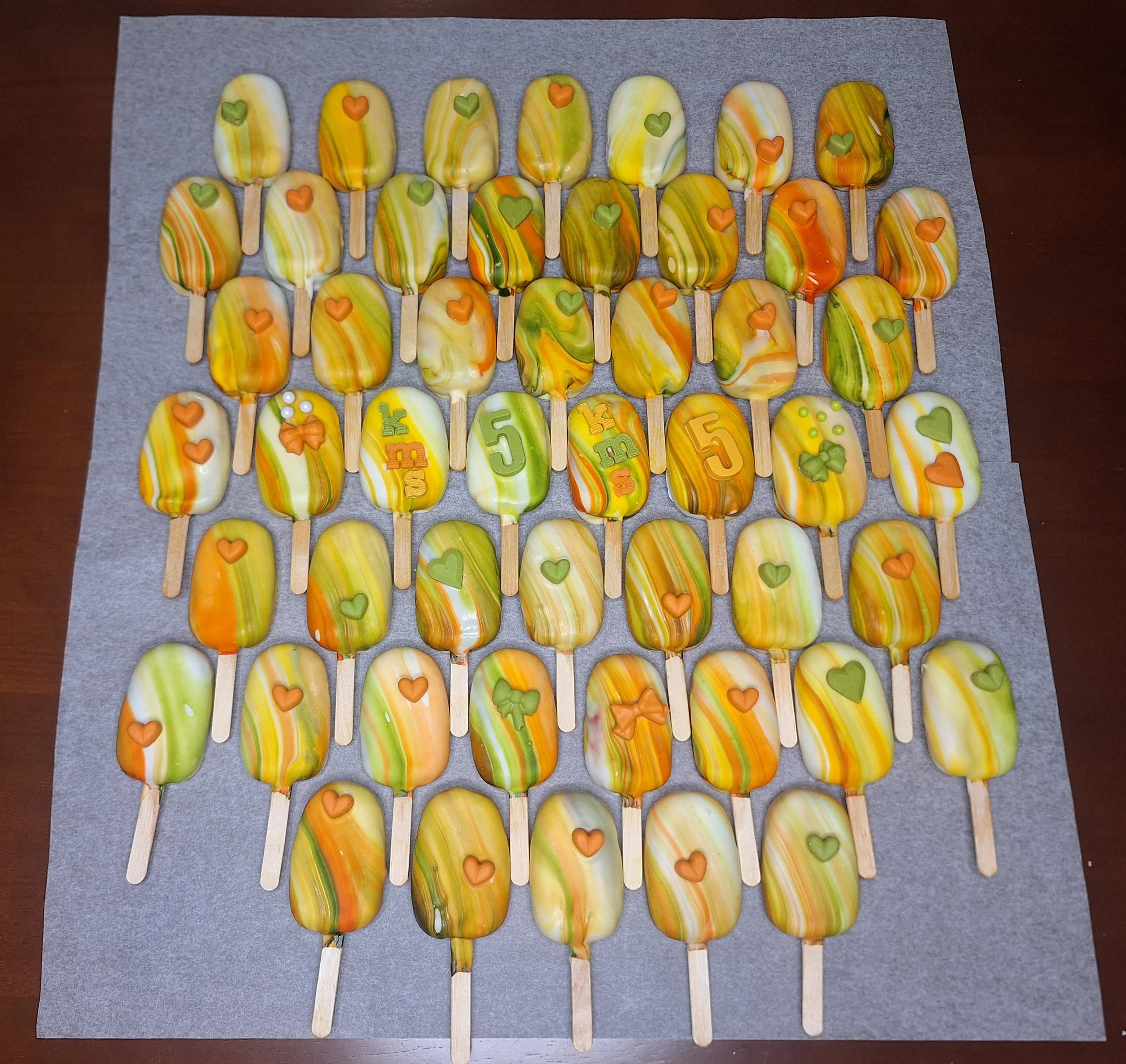 Colorful marble cakesicles arranged on parchment paper with chocolate shaped like hearts and numbers, along with additional decorations.