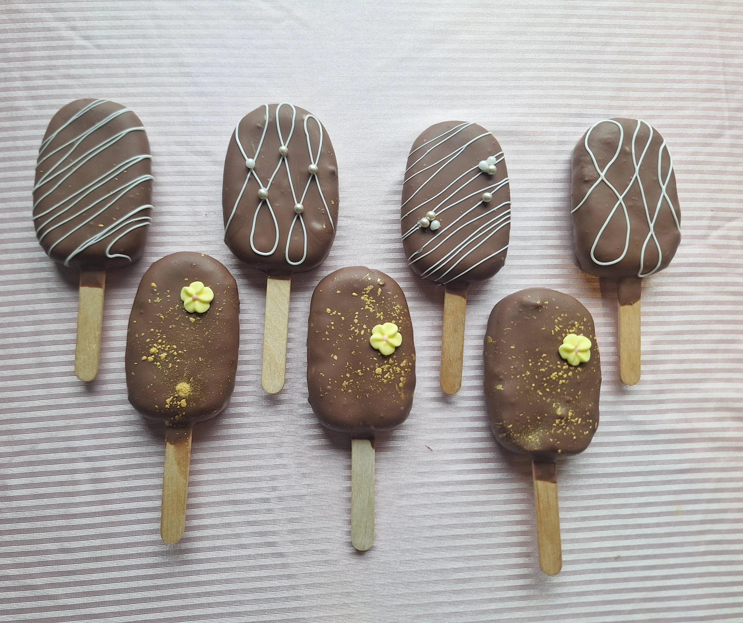 Chocolate-covered cakesicles on sticks decorated with white drizzle and small yellow flowers, placed on a pink striped background.