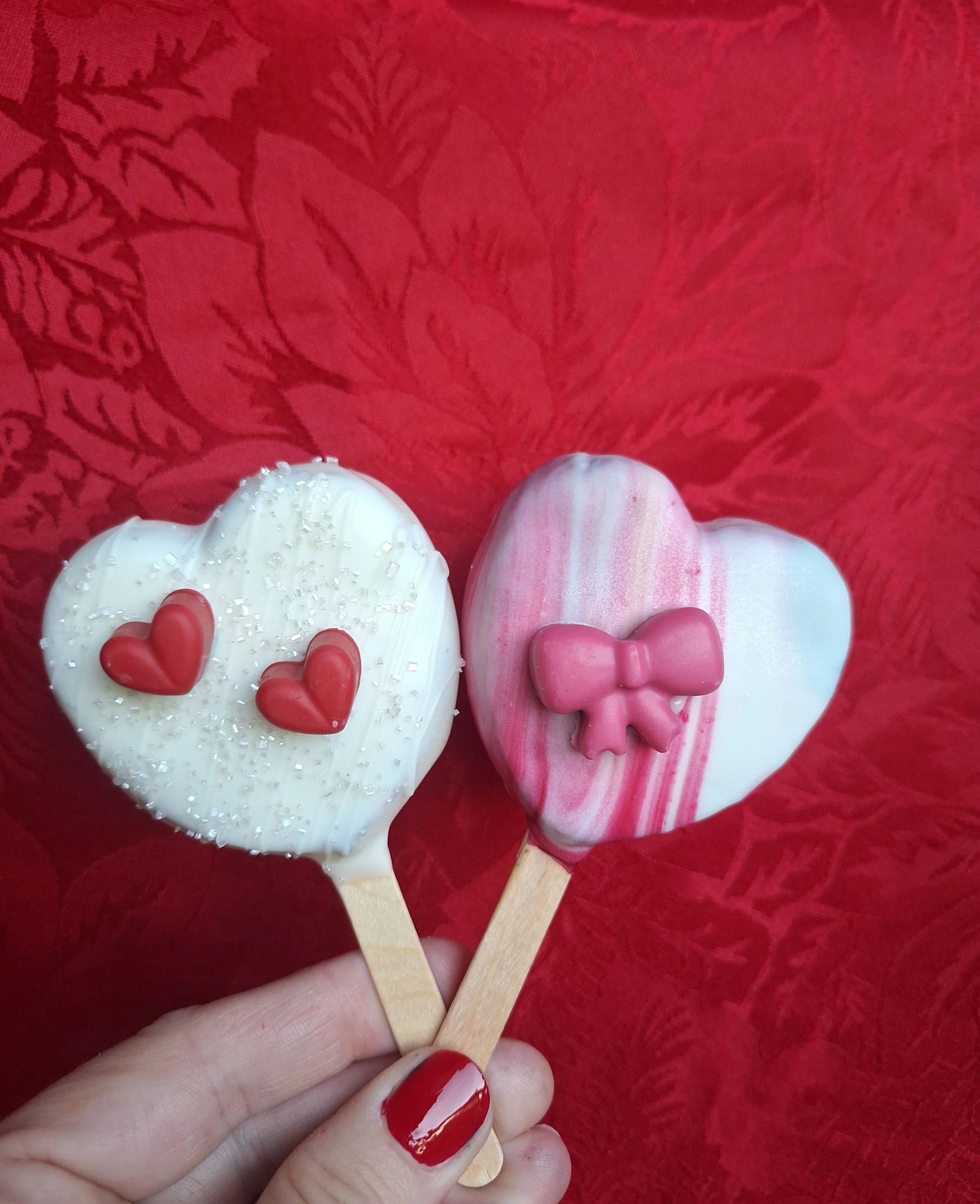 Close-up of a person's hand holding two Valentine's Day heart-shaped cake pops with red and pink decorations, resting on a red textured fabric background.