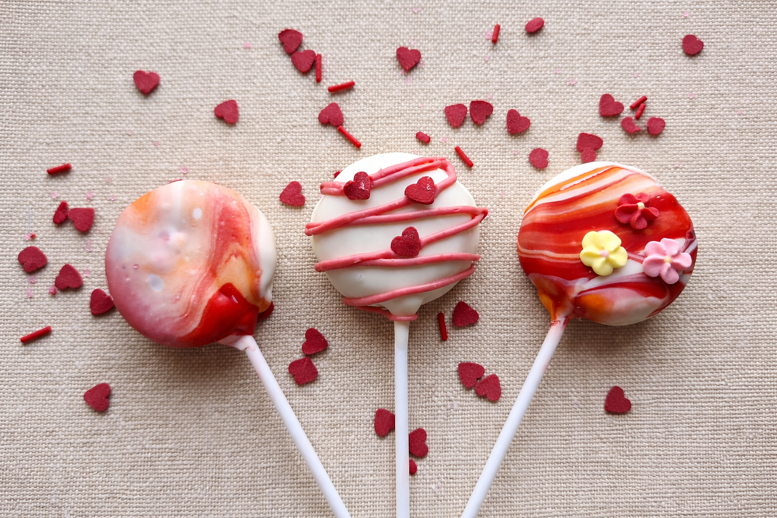 Three heart-shaped cake pops decorated with red, white, and pink chocolate and sprinkles, lying on a beige fabric surface surrounded by small red heart-shaped sprinkles.