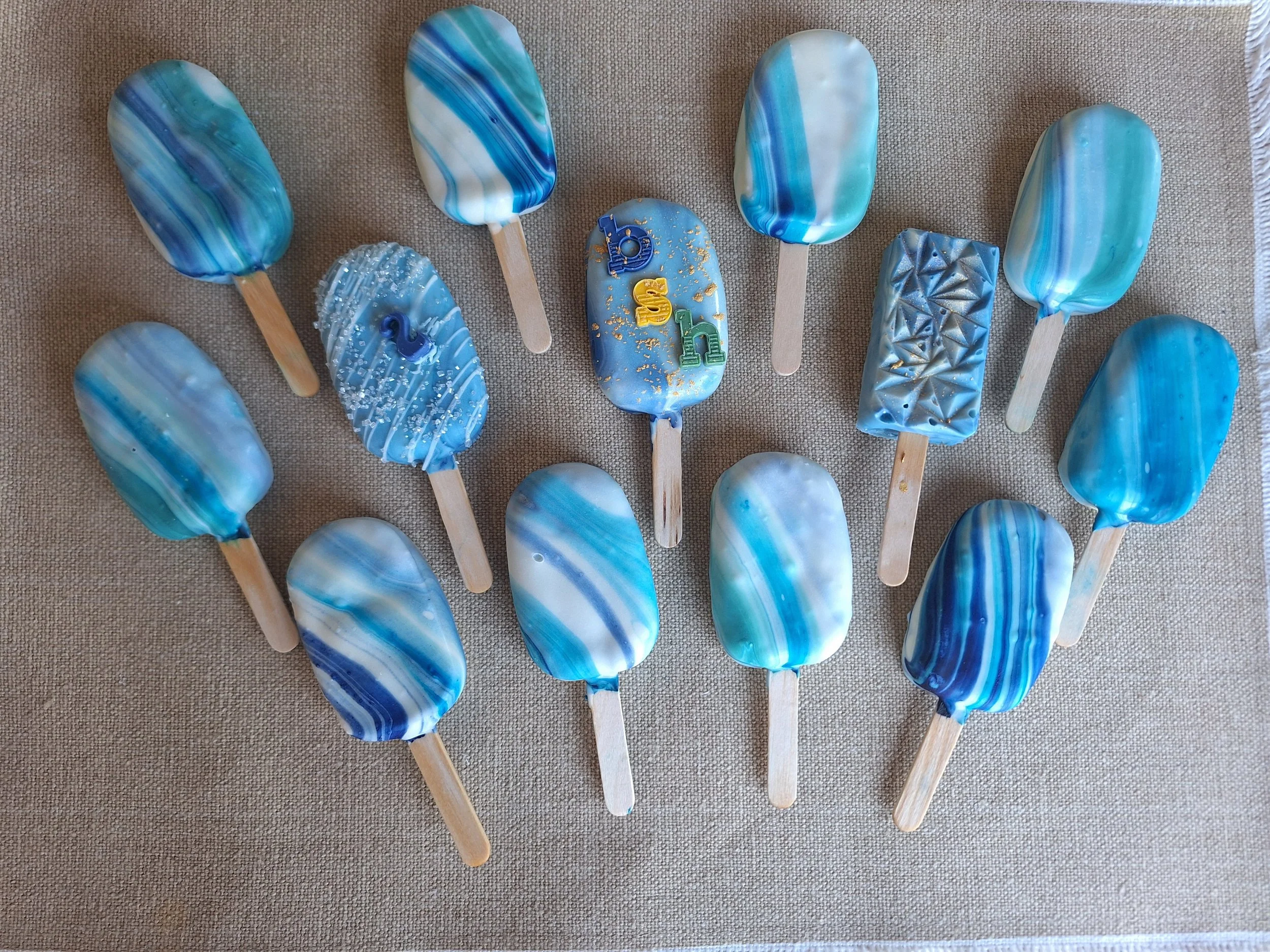 A collection of blue and white marbled cakesicles with wooden sticks, some decorated with sprinkles or gold flakes, arranged on a beige fabric surface.