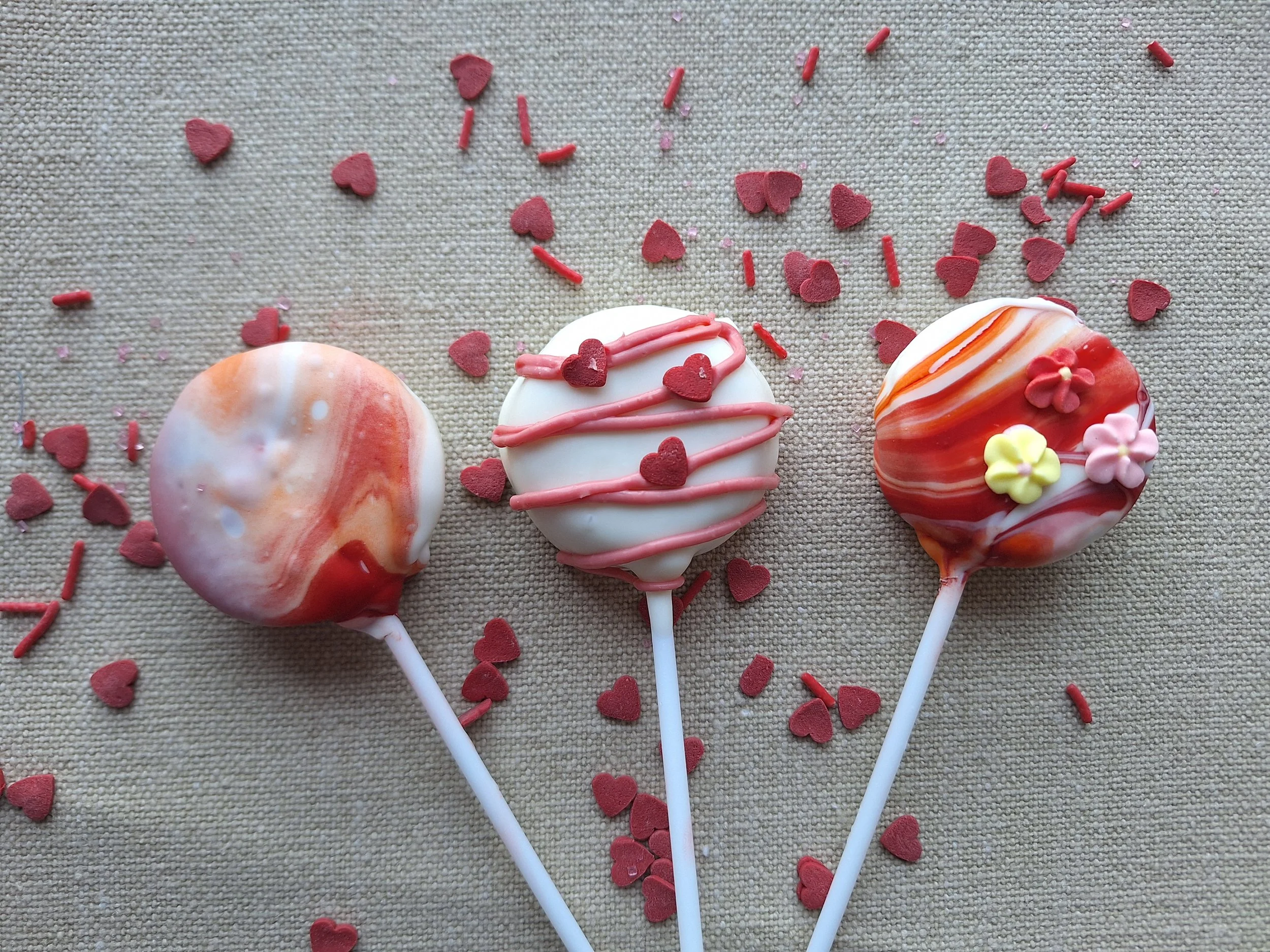 Colorful cake pops decorated with red, yellow, and pink flower and heart-shaped sprinkles on a textured beige surface.