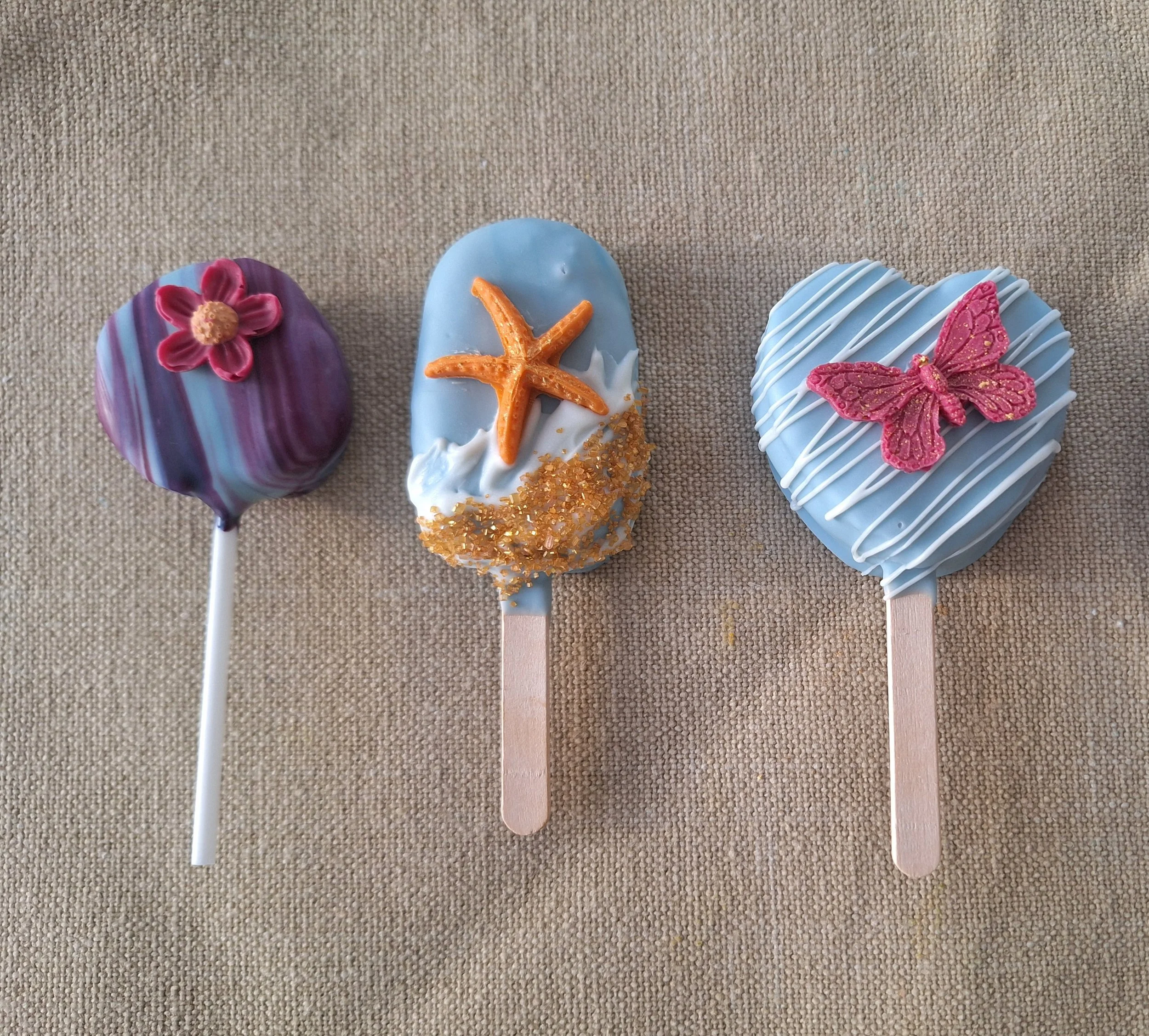 Three decorated cakesicles on a textured beige surface.