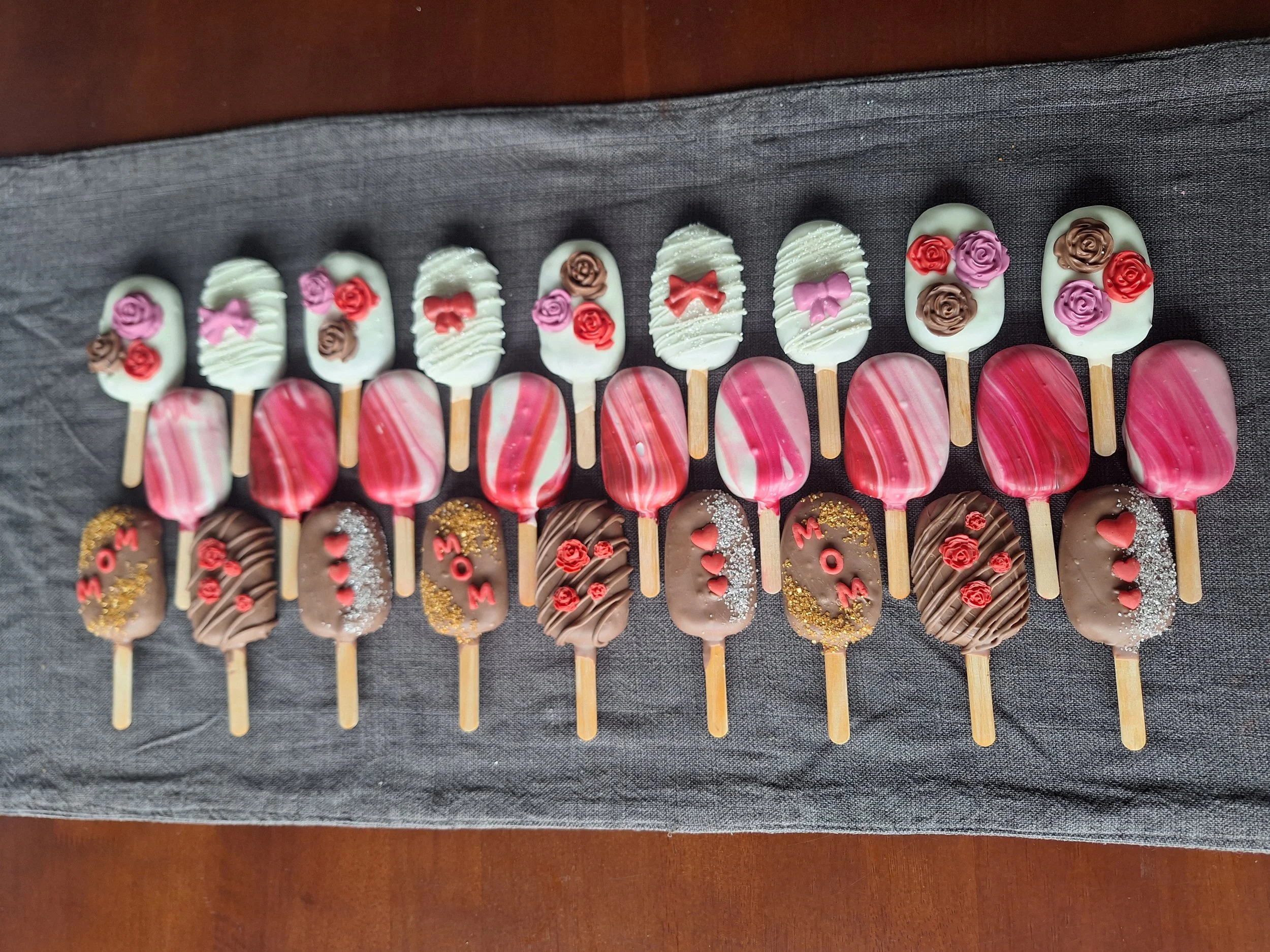 Assorted decorated cakesicles arranged in a row on black fabric, featuring various colors and designs including roses, bows, and patterns.