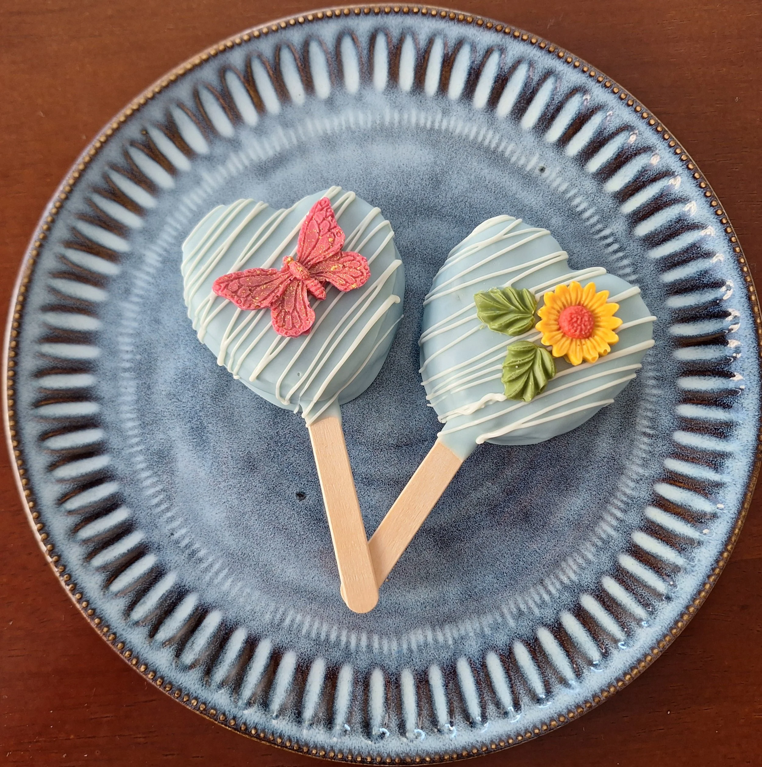 Decorated cake pops on a blue ceramic plate, including a cake pop with a pink butterfly, a sunflower, and green leaves, on a brown wooden surface.