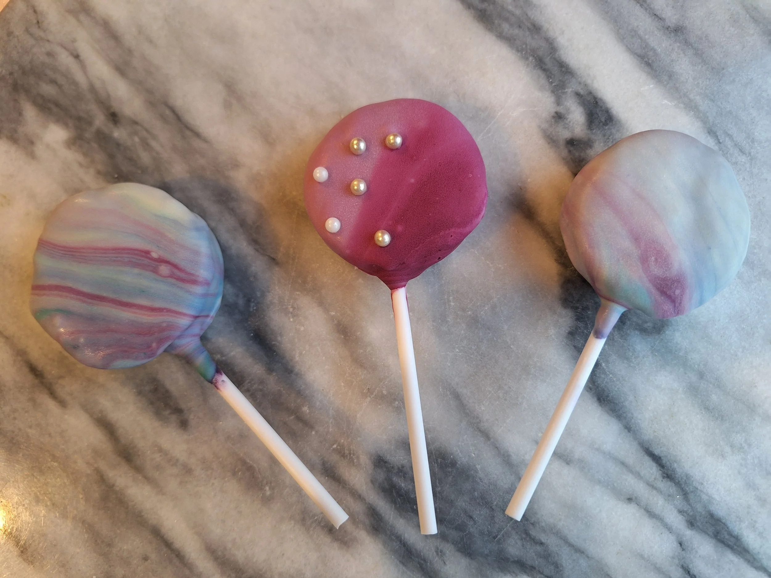 Three colorful swirled cake pops with pearl-like decorative beads on a marble surface.
