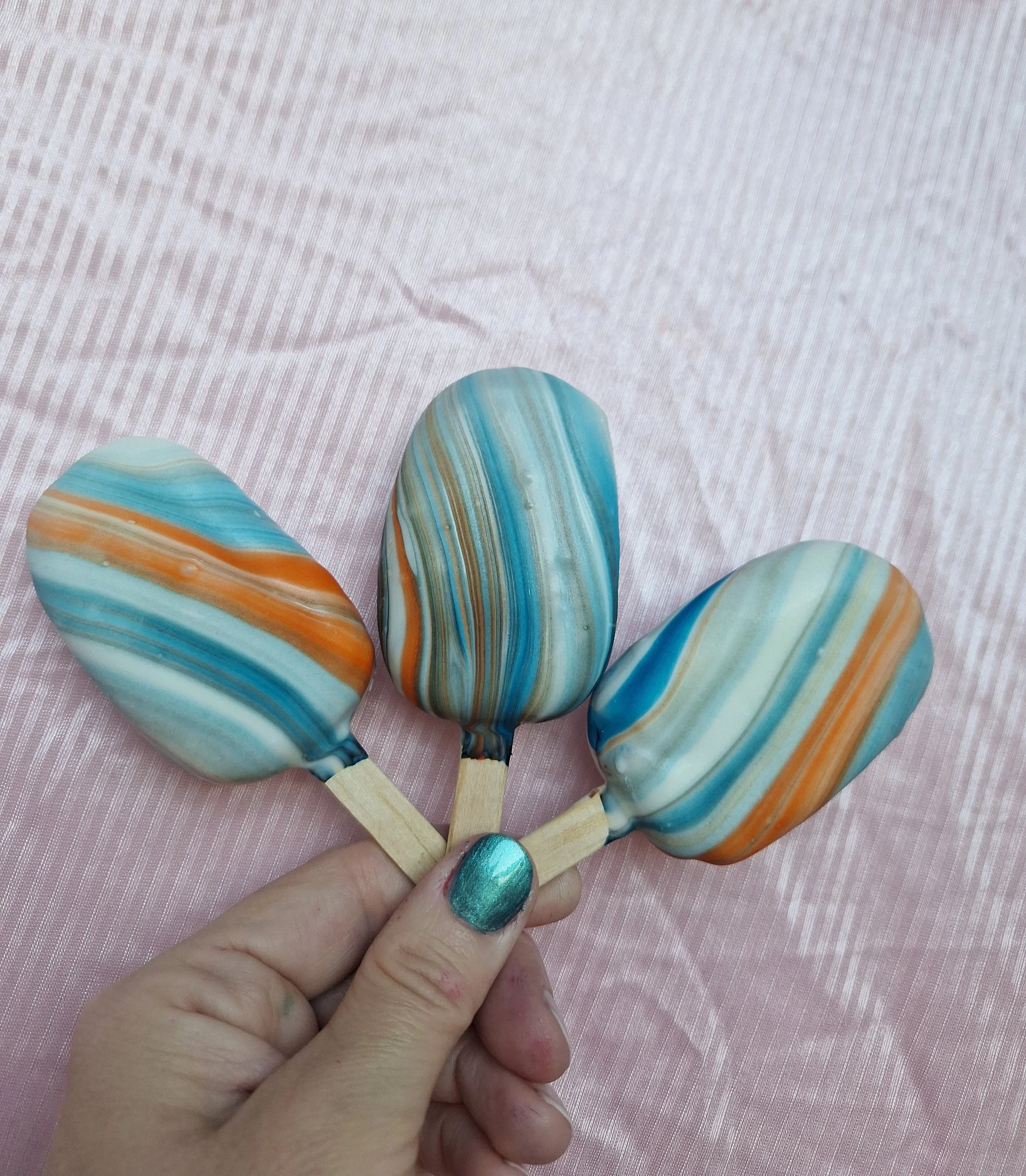 Colorful marbled cakesicles held by a person with teal nail polish, resting on a pink textured surface.