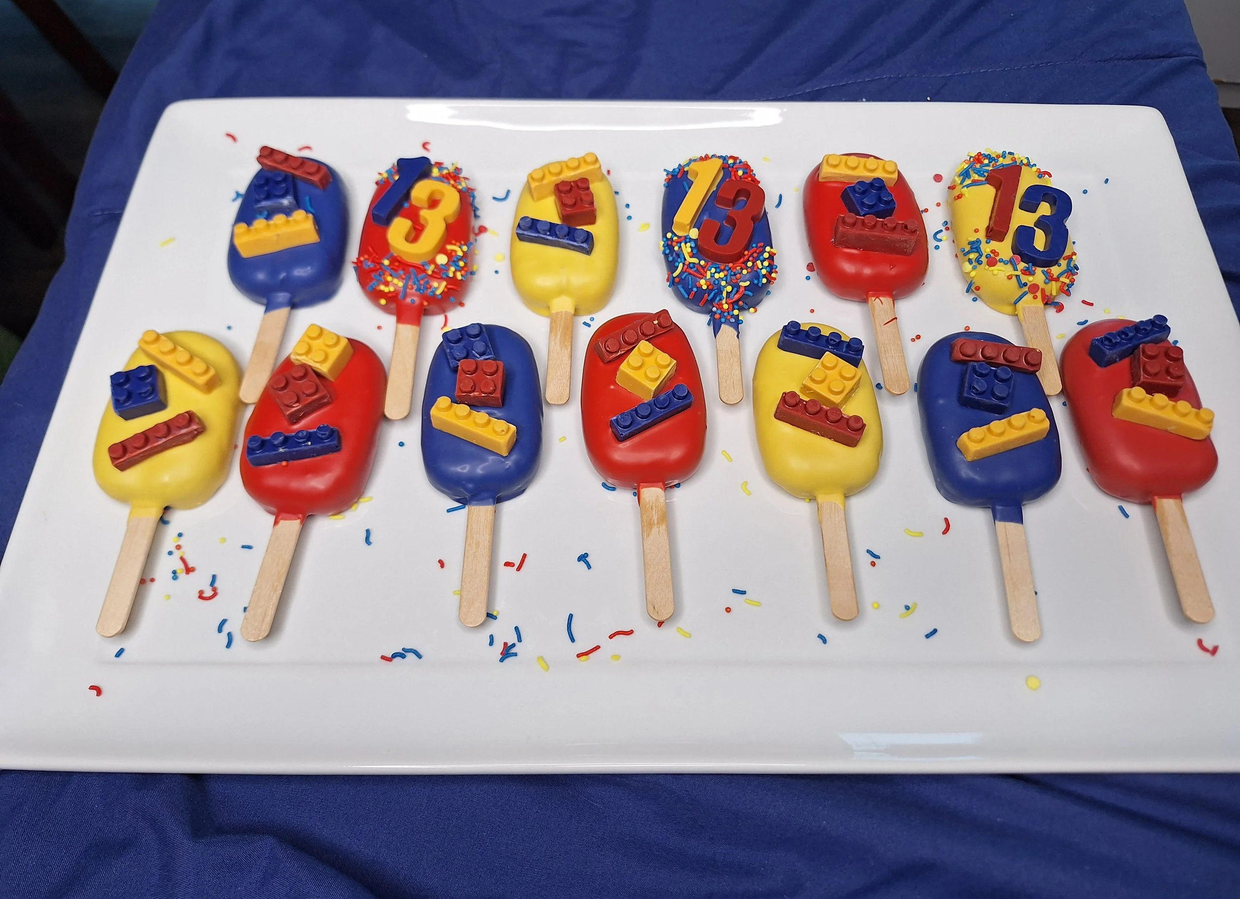 Colorful cake pops decorated with blue, red, and yellow icing, topped with Lego brick-shaped chocolate, arranged on a white tray with colorful confetti scattered around.