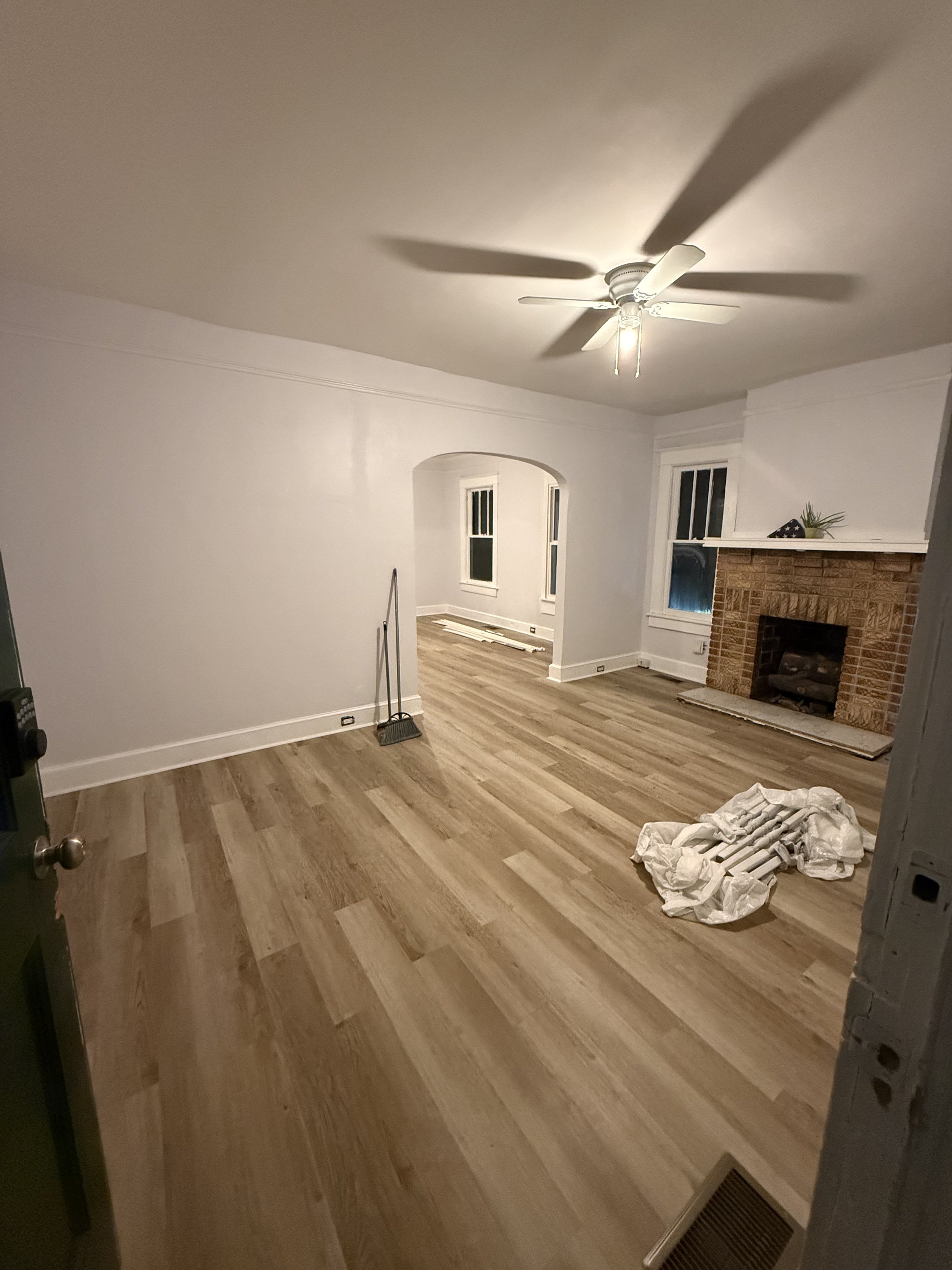 A living room with light-colored walls, hardwood flooring, a white ceiling fan with lights, and a brick fireplace. There is a small plant and a folded American flag on the mantel. Some materials and tools are on the floor, indicating ongoing renovation.