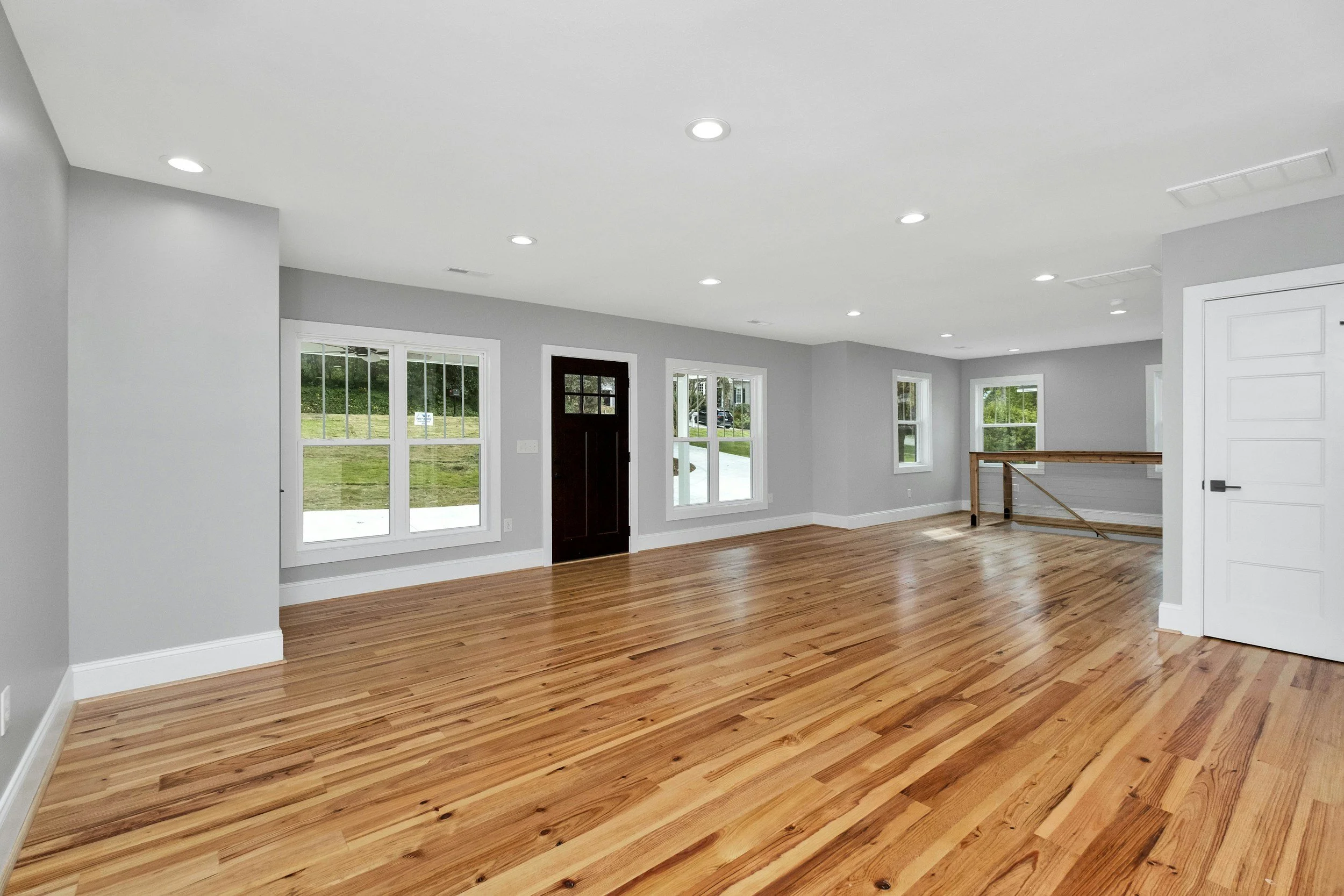 Interior of an empty living room with light gray walls, multiple large windows, hardwood floors, and ceiling lights.