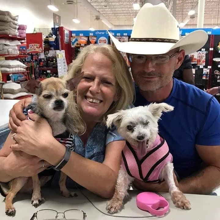 A smiling woman and man with two small dogs in a pet store, the woman holding one dog and the man wearing a cowboy hat.