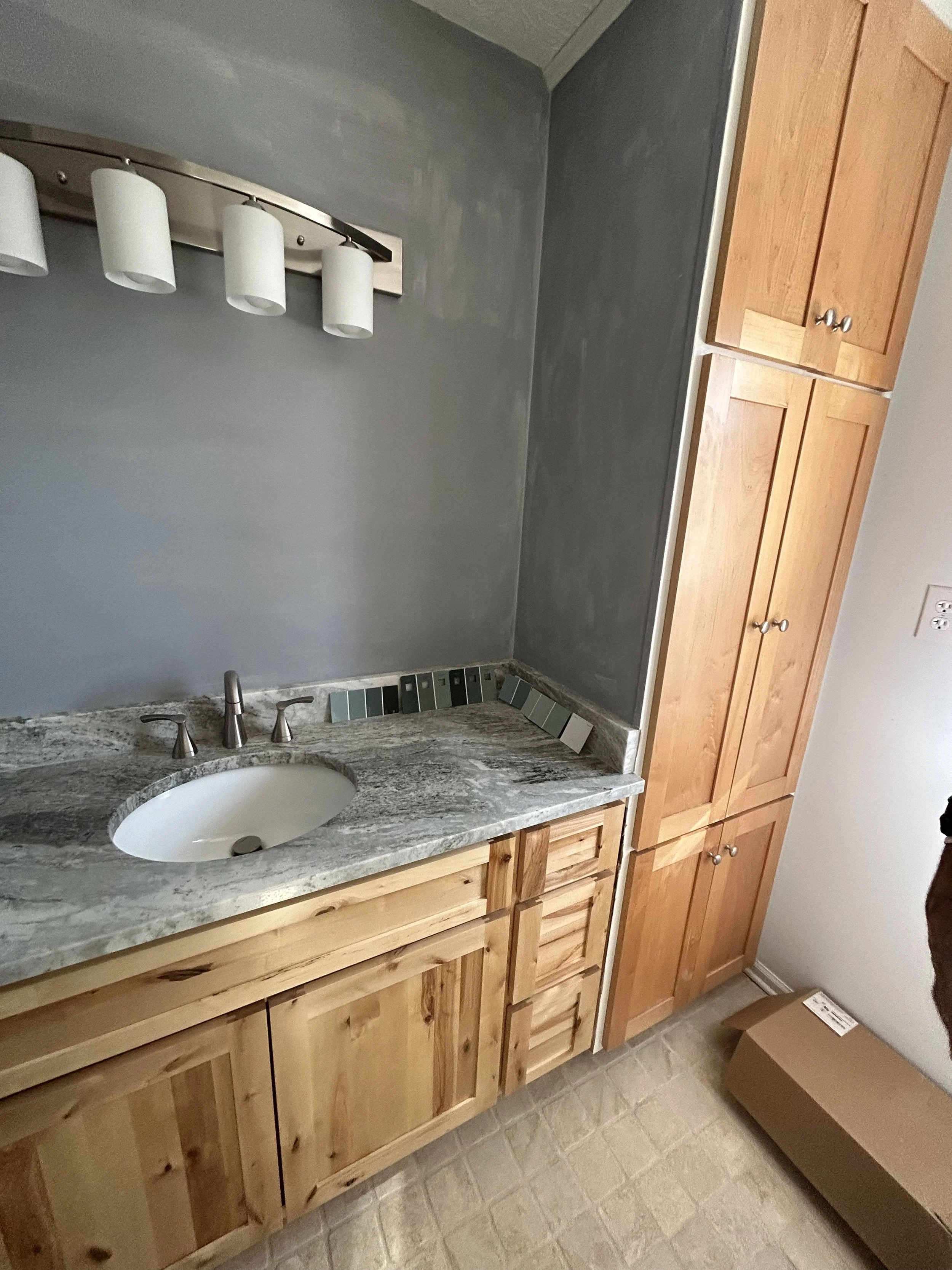 A bathroom with a gray painted wall, a wooden cabinet, a marble countertop with a built-in sink, a metallic faucet, a light fixture with four white cylindrical shades, and paint color samples along the edge of the countertop.