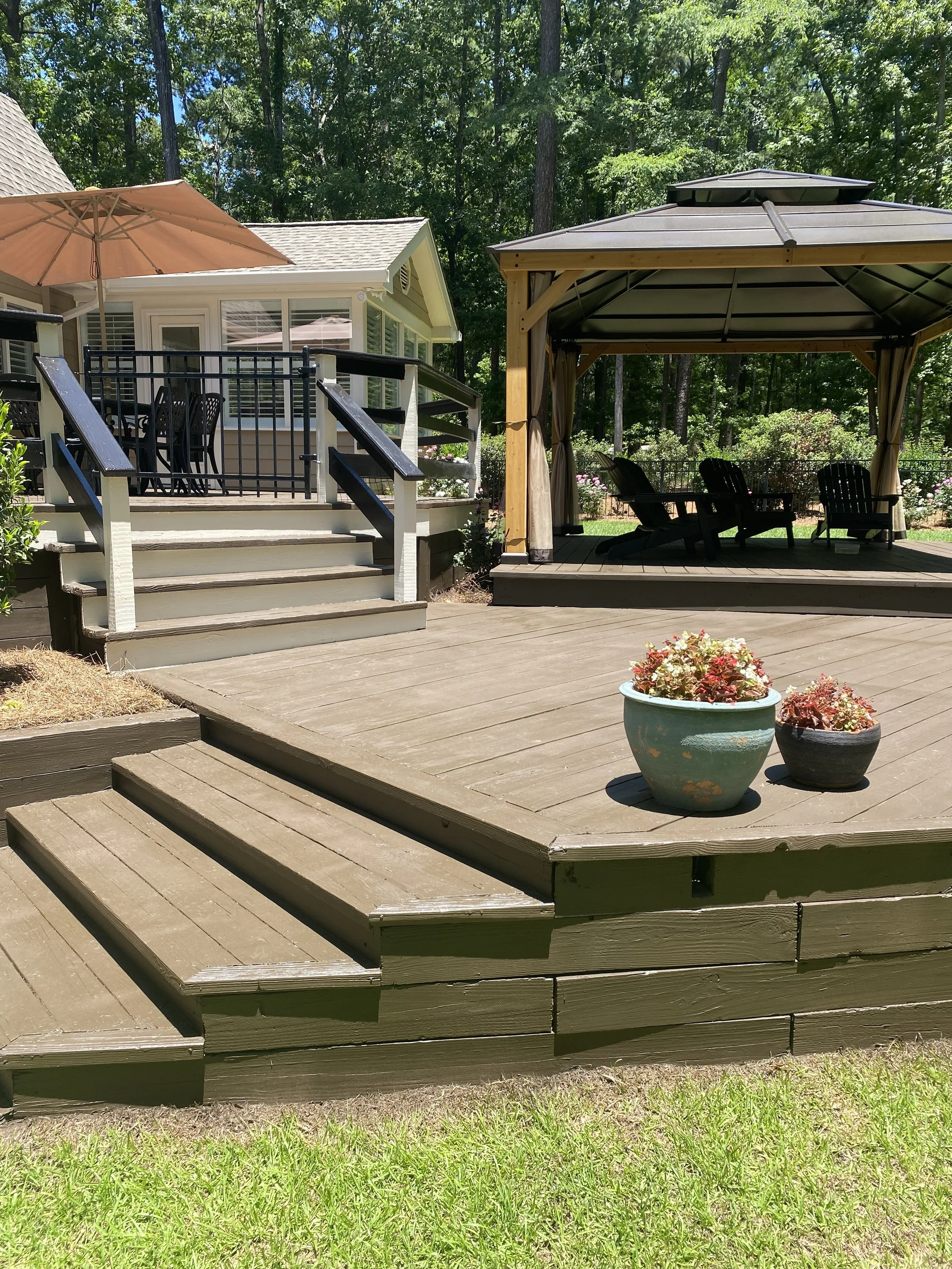 A wooden deck with stairs leading up to a house with a screened porch, and a gazebo with seating inside surrounded by trees, bushes, and potted plants.