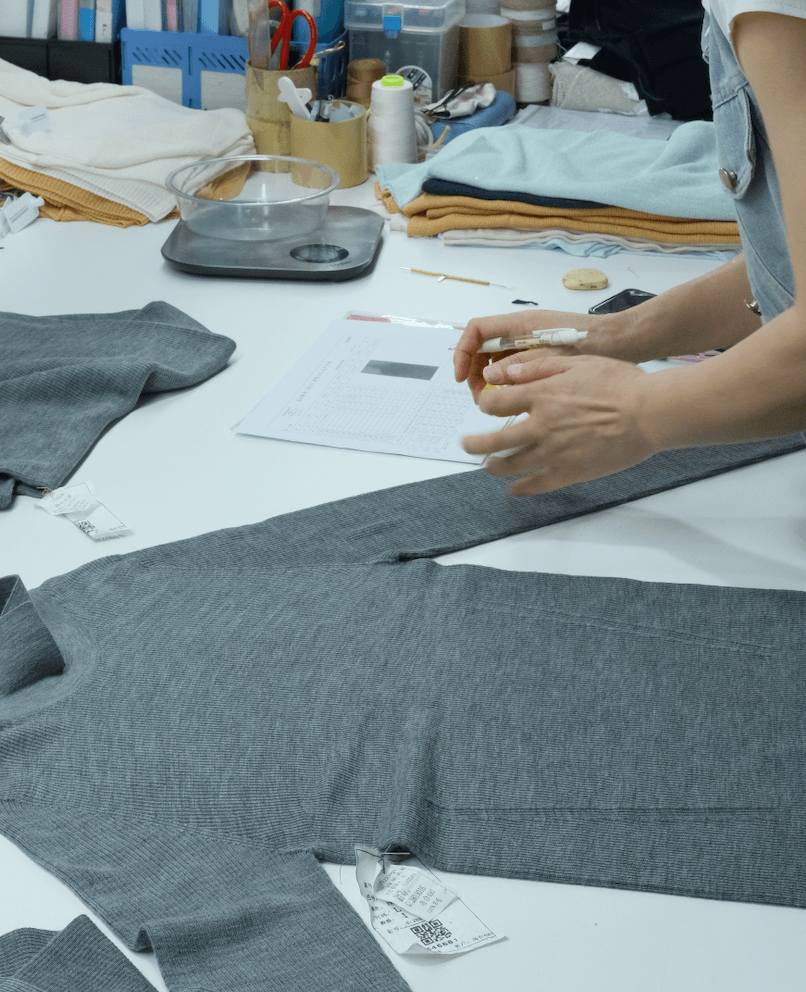 A grey knitted sweater sample being checked on a worktable with measurement notes, showing the quality control and sample review process.