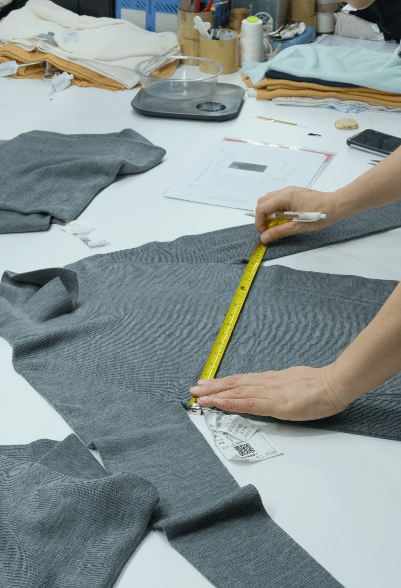 A grey knitwear sample being measured against a spec sheet, showing sample review and quality control checks before production approval.