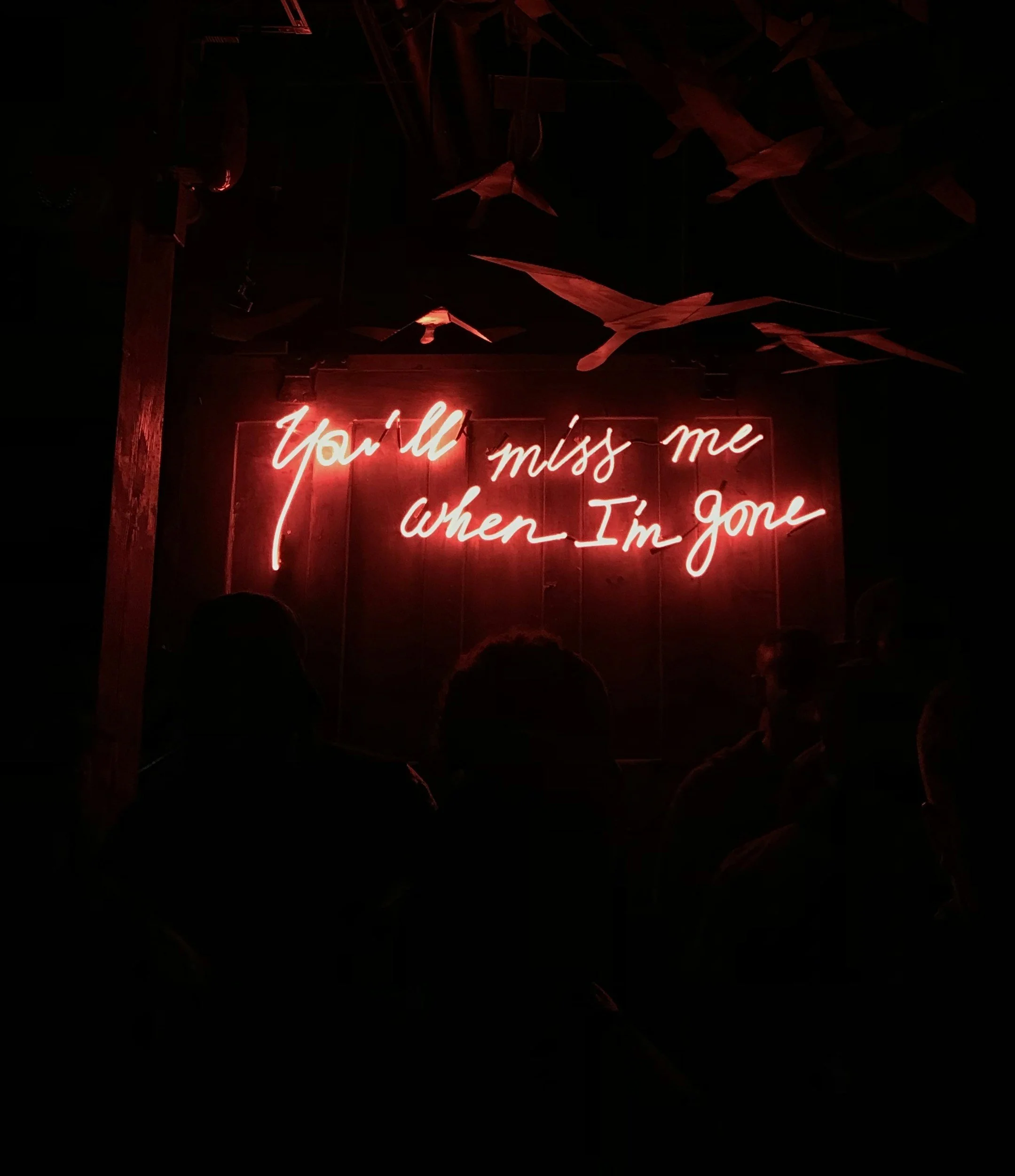 Neon sign with the phrase "You’ll miss me when I’m gone" in red, surrounded by origami cranes hanging from the ceiling.