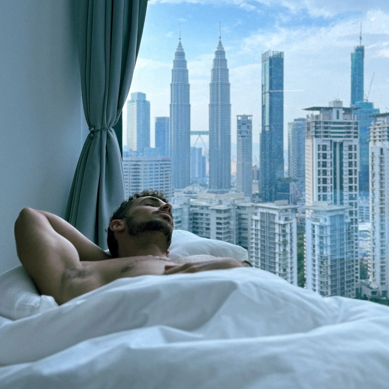 A shirtless man with a beard and short hair lies in bed with his eyes closed, resting with his arms behind his head, in a high-rise apartment overlooking a city skyline with tall buildings, including the Petronas Towers in Kuala Lumpur.