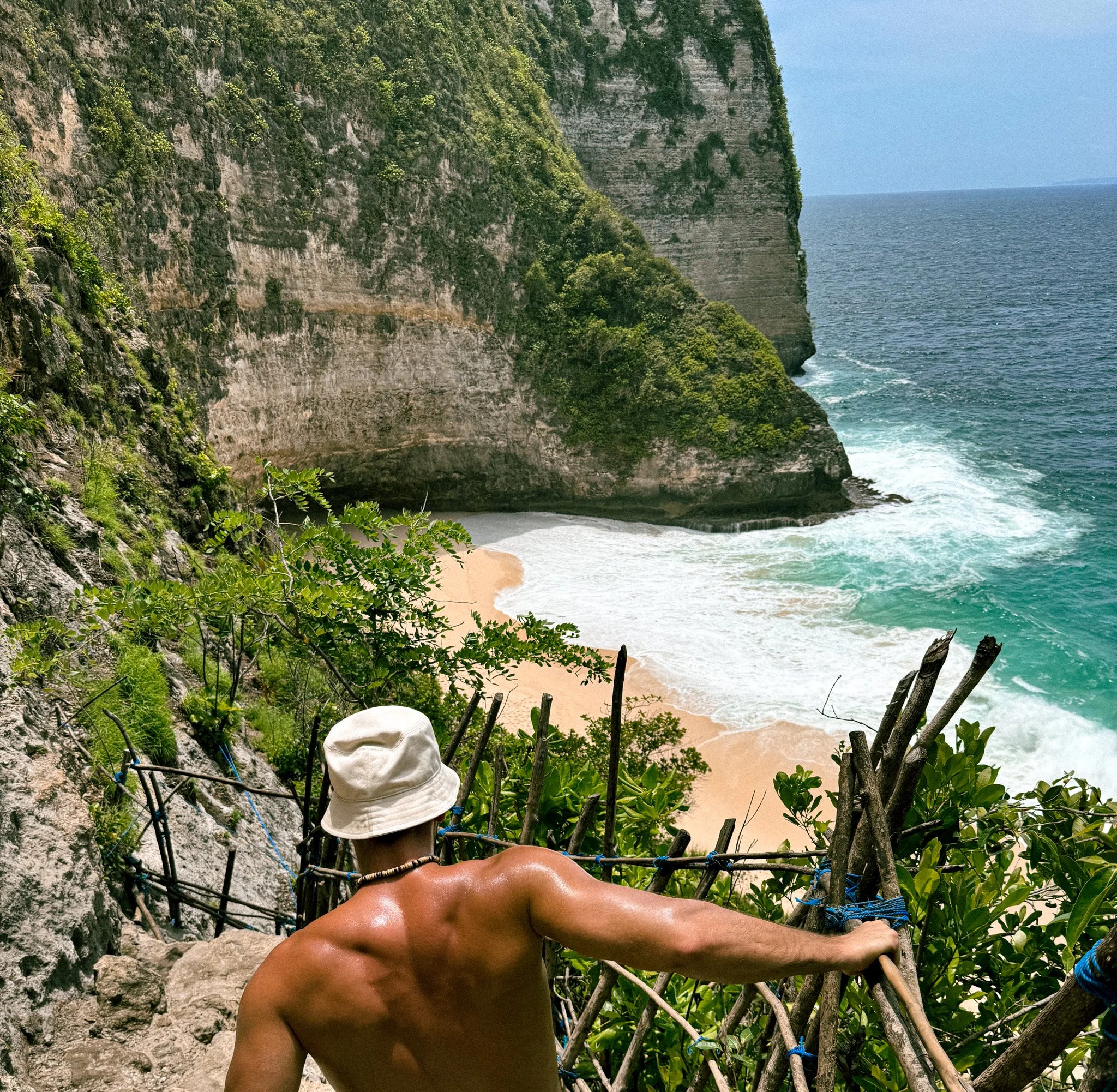 A shirtless person with a white hat climbing a steep rock face surrounded by green plants, overlooking an ocean with cliffs and blue sky.