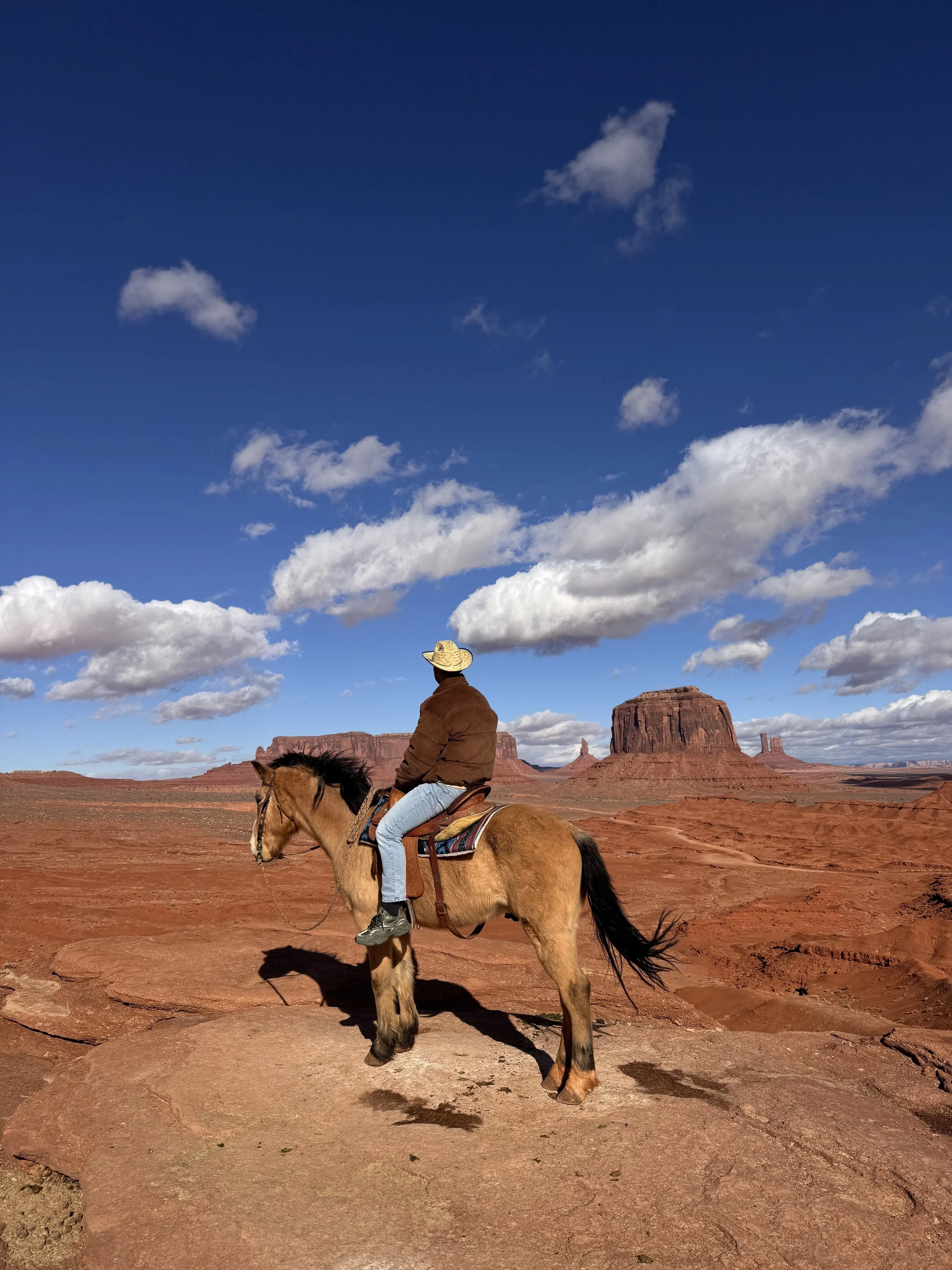 A person riding a horse in a desert landscape with red rock formations and a partly cloudy sky.