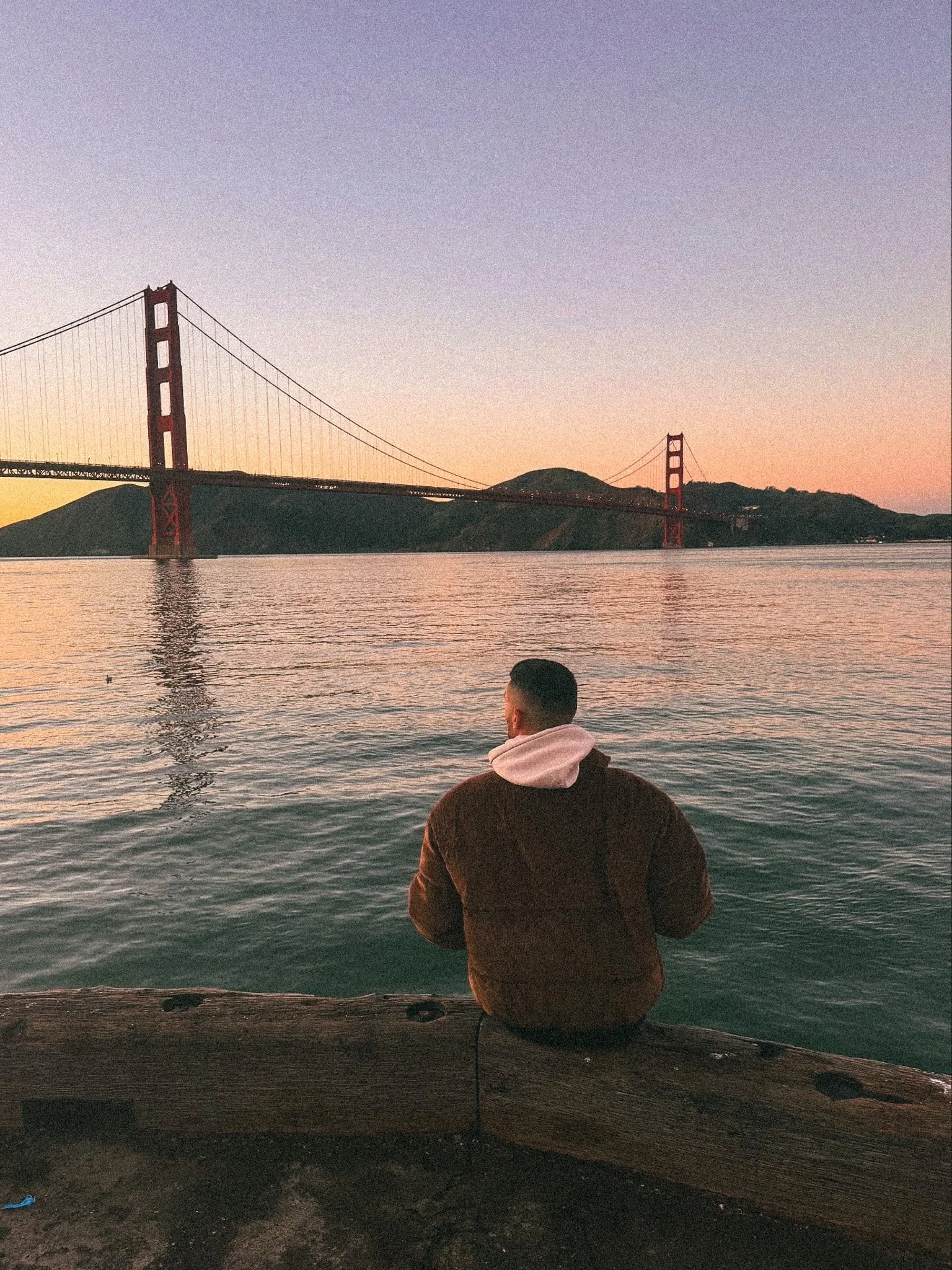 A person with short hair and wearing a brown hoodie, sitting on a dock, looking at the water during sunset, with the Golden Gate Bridge in the background.