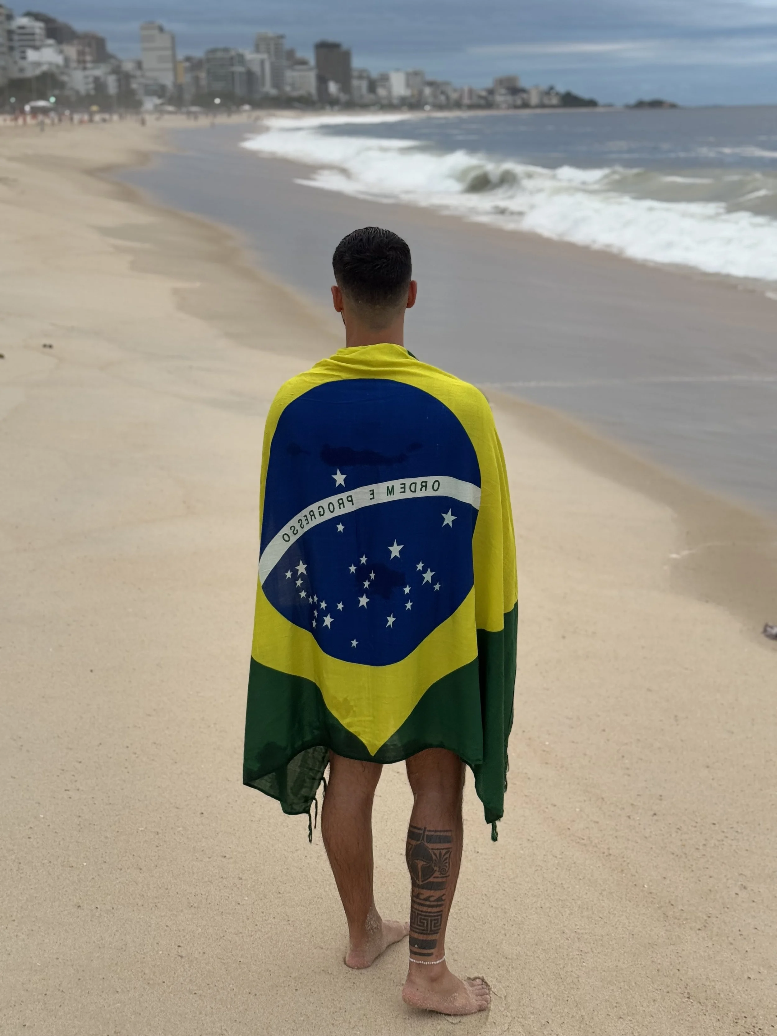 A person standing on a sandy beach, wrapped in a Brazilian flag, facing the ocean with waves. In the background, there is a city skyline with tall buildings and a cloudy sky.