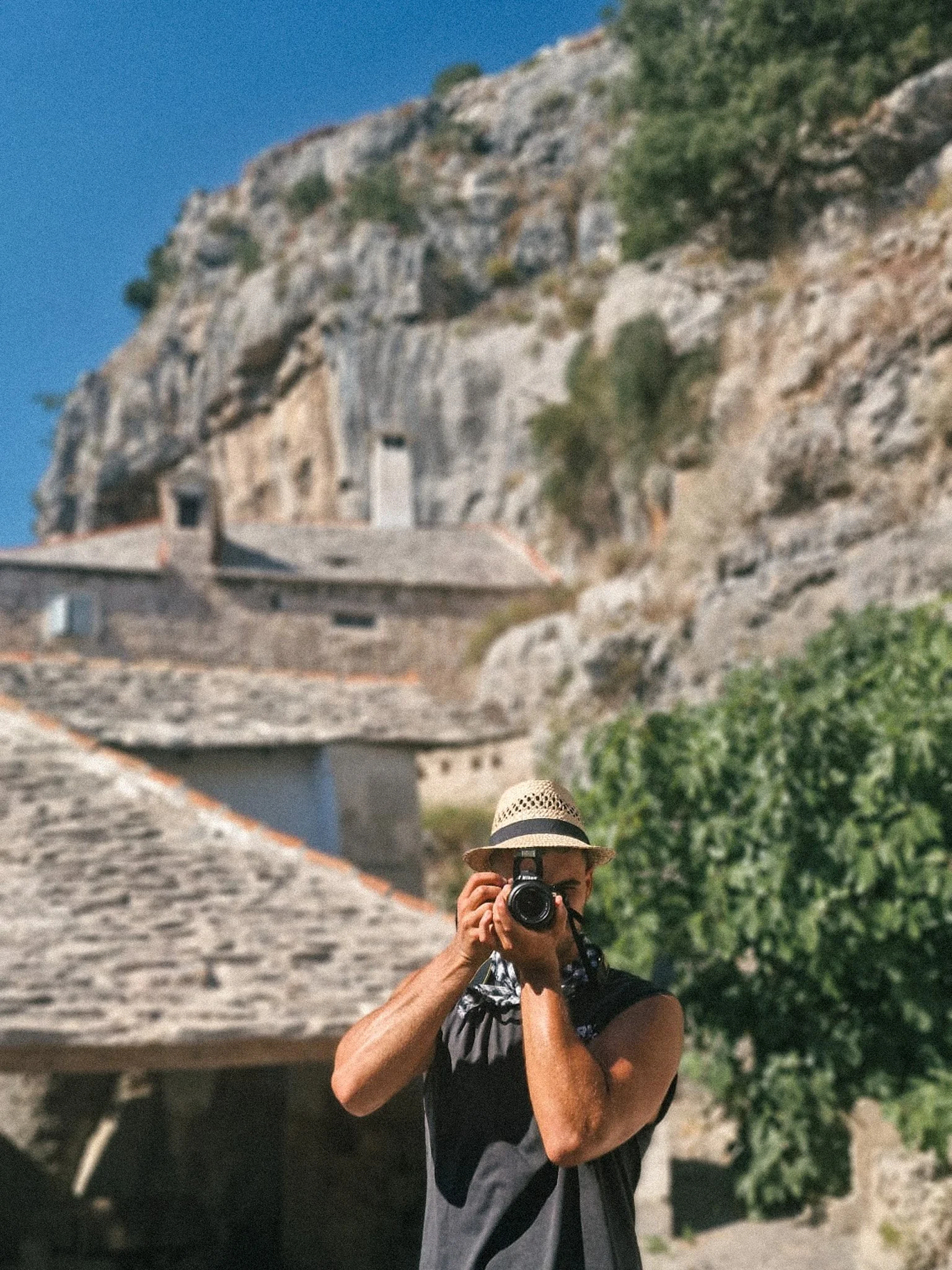 Person wearing a hat taking a photograph with a camera in front of a rocky hillside with greenery and buildings.