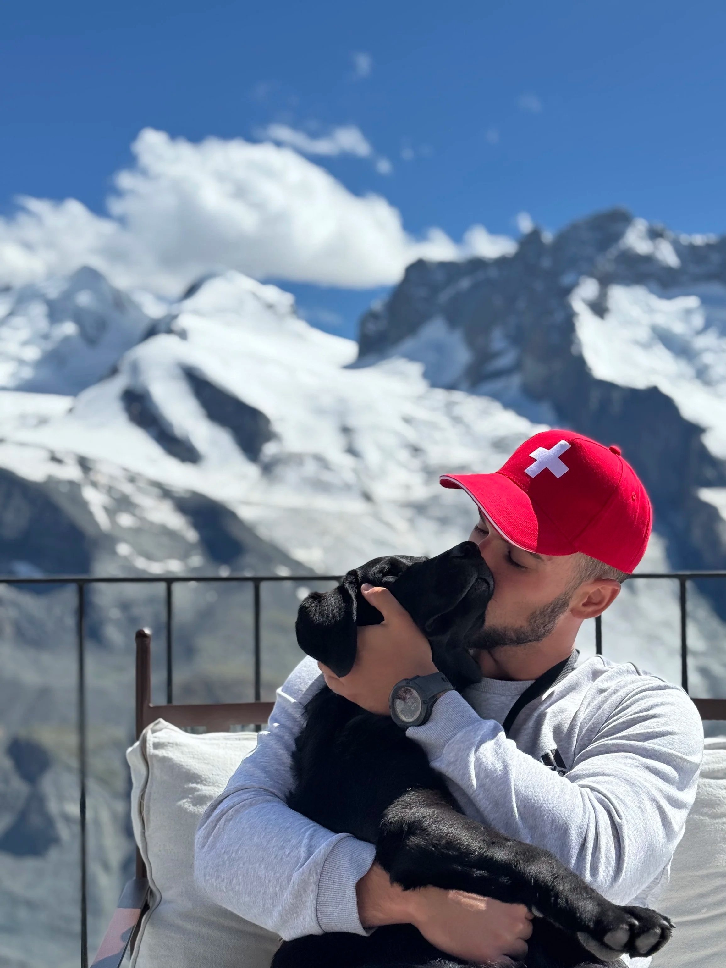 A man wearing a gray jacket and a red cap with a white cross is holding a black puppy in his arms with a mountain landscape in the background.