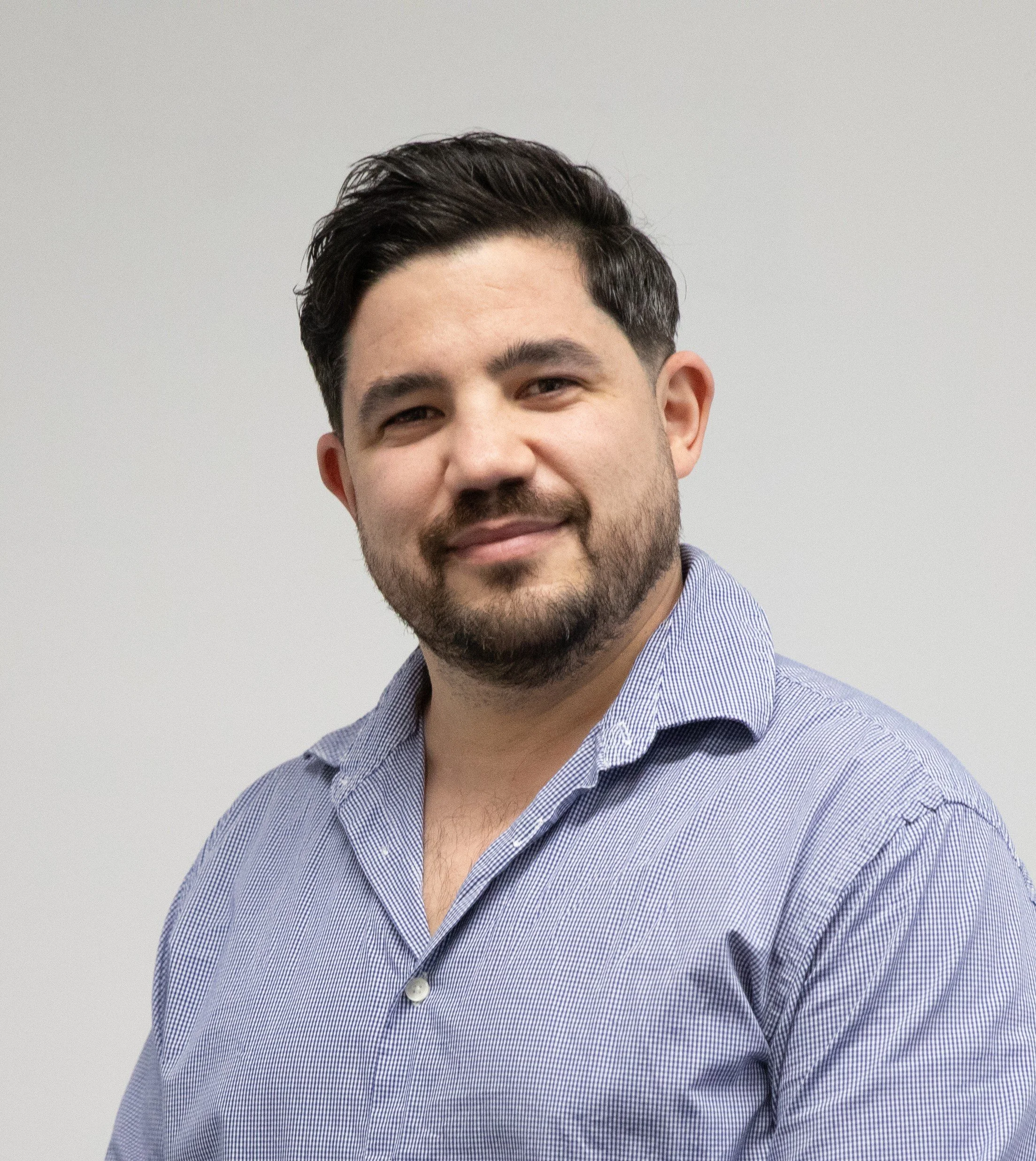 Close-up of a smiling man with dark, styled hair, a beard, and wearing a blue and white checkered shirt against a plain gray background.