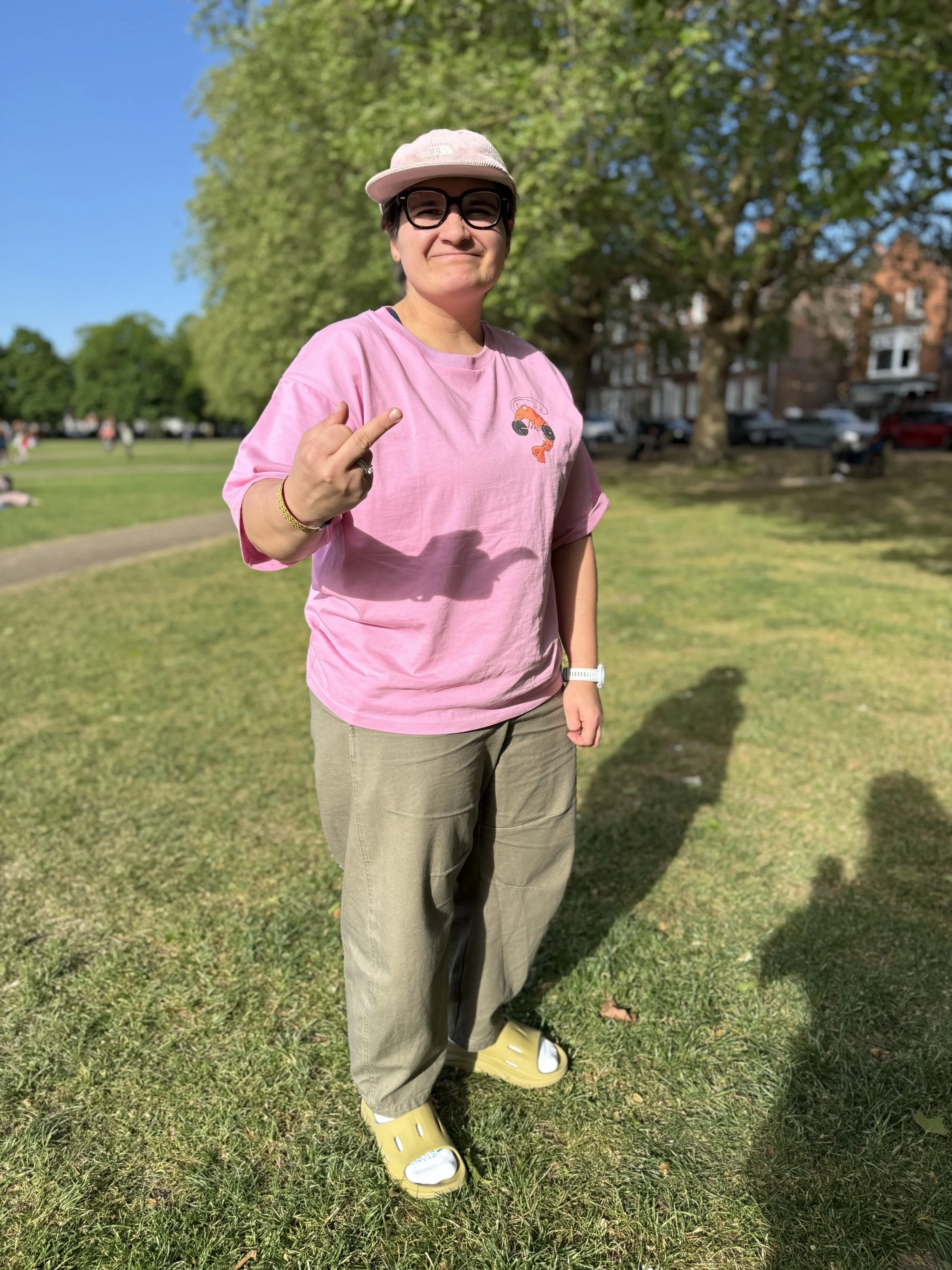 Yasmin standing on Richmond Green, wearing the oversized pink Mr Prawn tee and raising her middle finger as her love language.