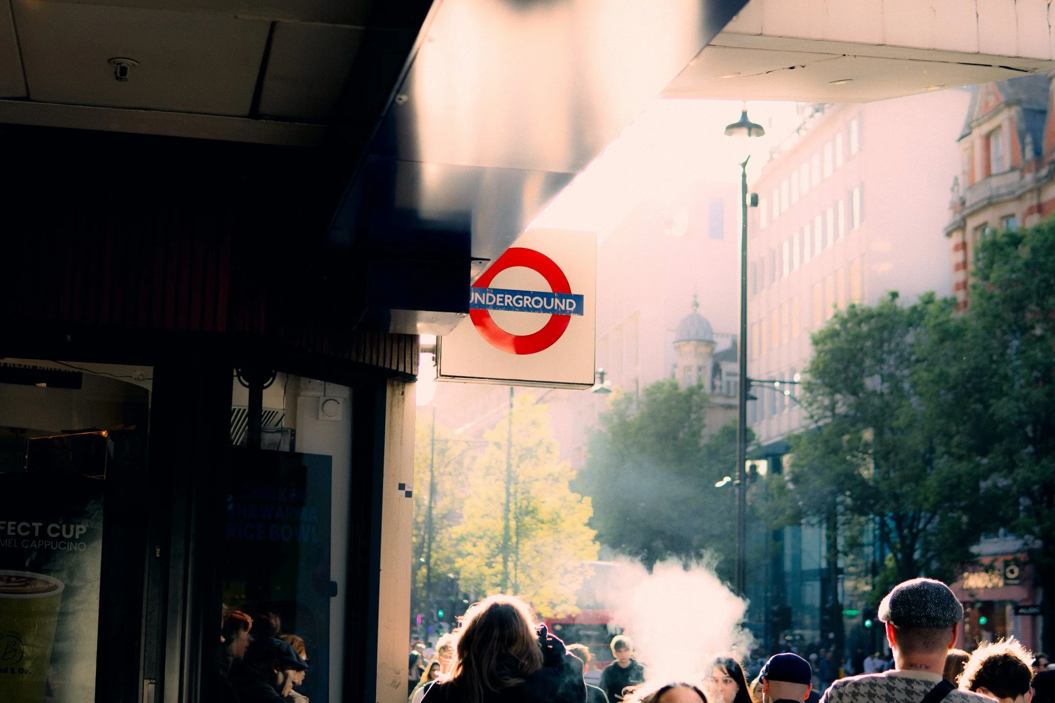 Street scene with a crowd of people near a subway station entrance, with a red and blue 'Underground' sign, sunlight creating a hazy atmosphere, and buildings in the background.