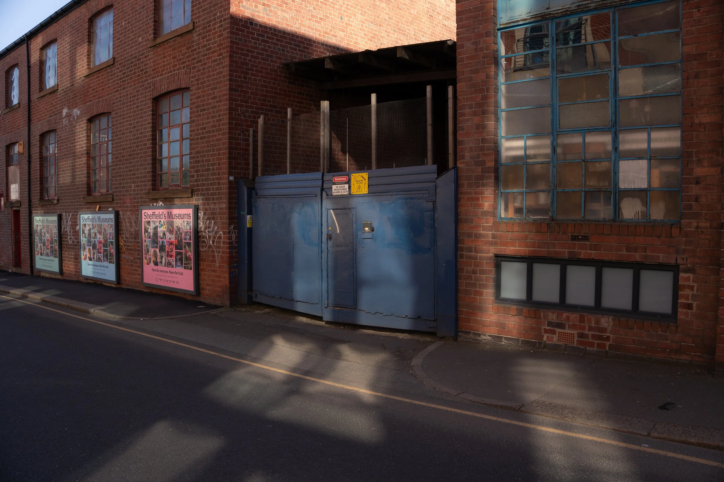 View of a brick building with multiple windows, some covered with metal panels. There are posters on the lower part of the building advertising Sheffield's Museums. A large blue gate and some warning sign are also visible.