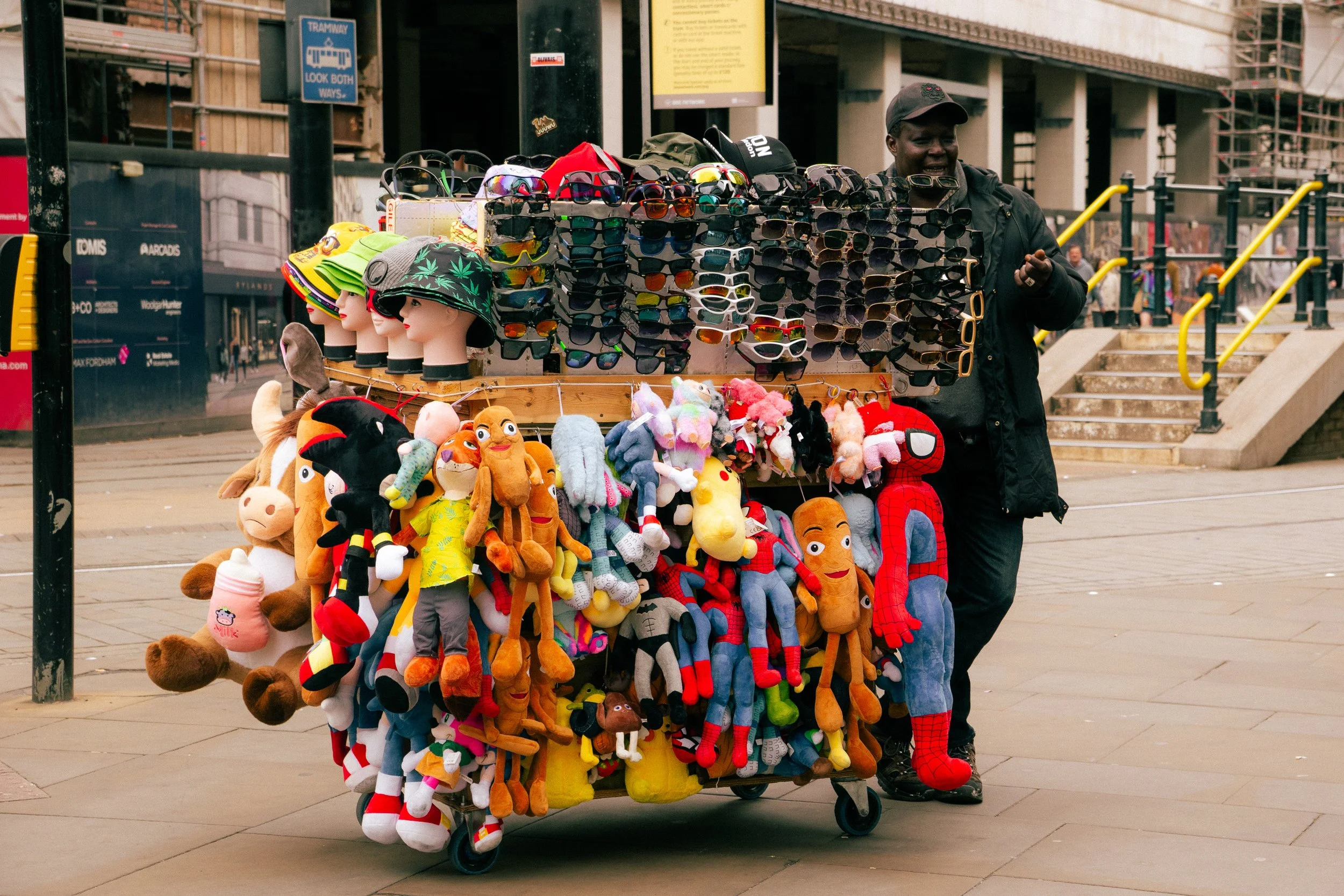 A street vendor’s cart displays sunglasses, hats, plush toys, and stuffed animals. A man stands behind the cart, partially obscured by the items. The background shows city stairs and buildings.